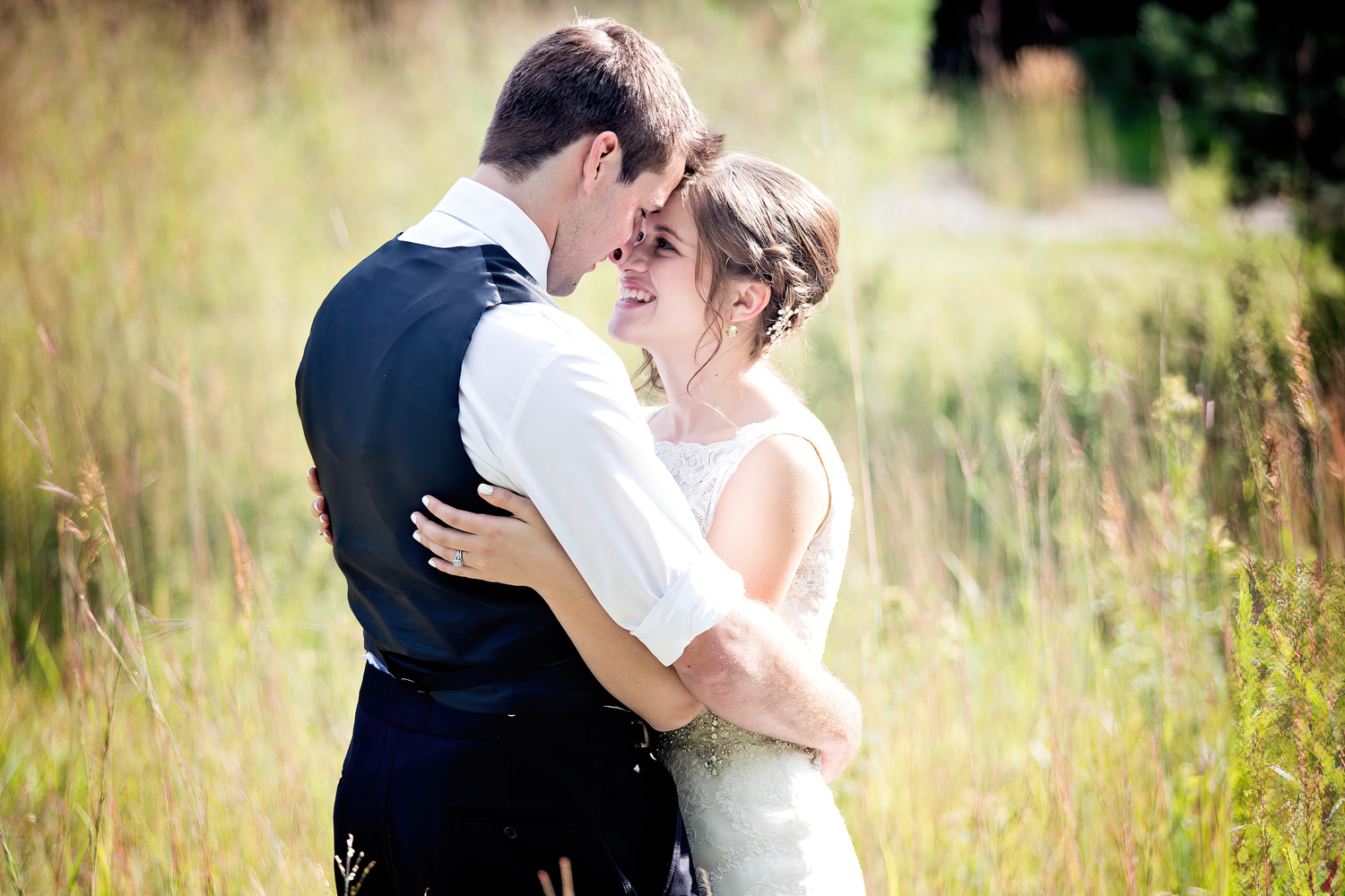 A romantic couple embraces in a sunlit meadow with tall grass during a wedding photo session.