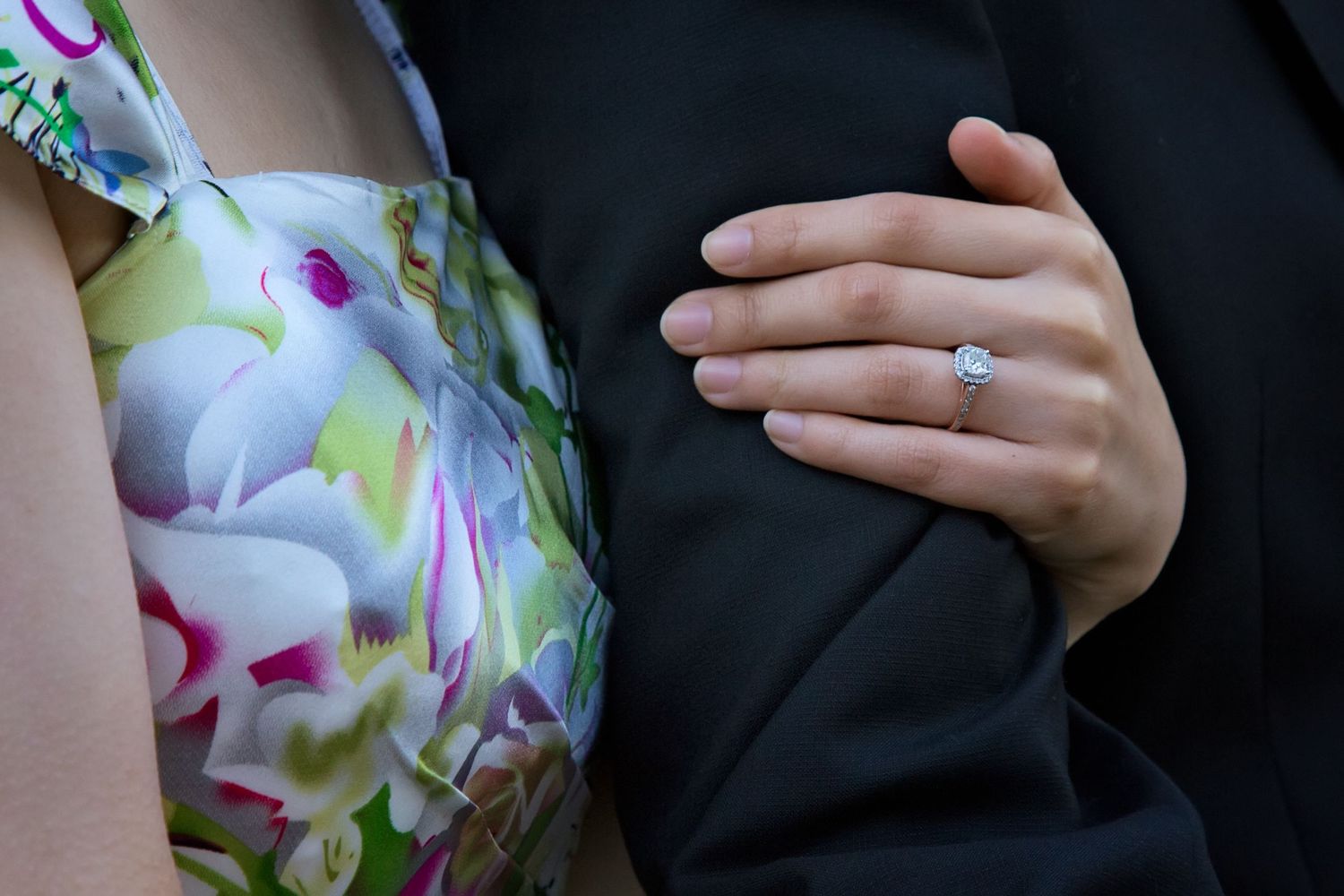 Close-up of hands embracing with engagement ring visible against floral dress and dark suit.