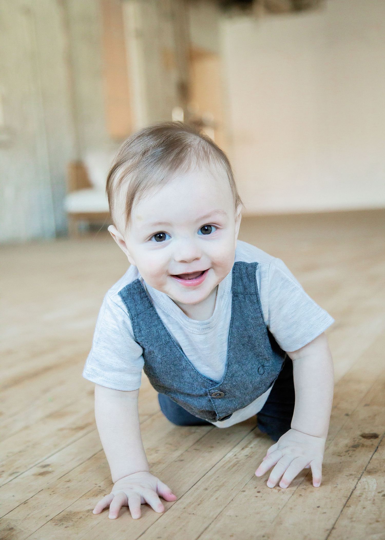 Baby in a gray vest and white shirt crawls on wooden floor with a bright, cheerful expression.