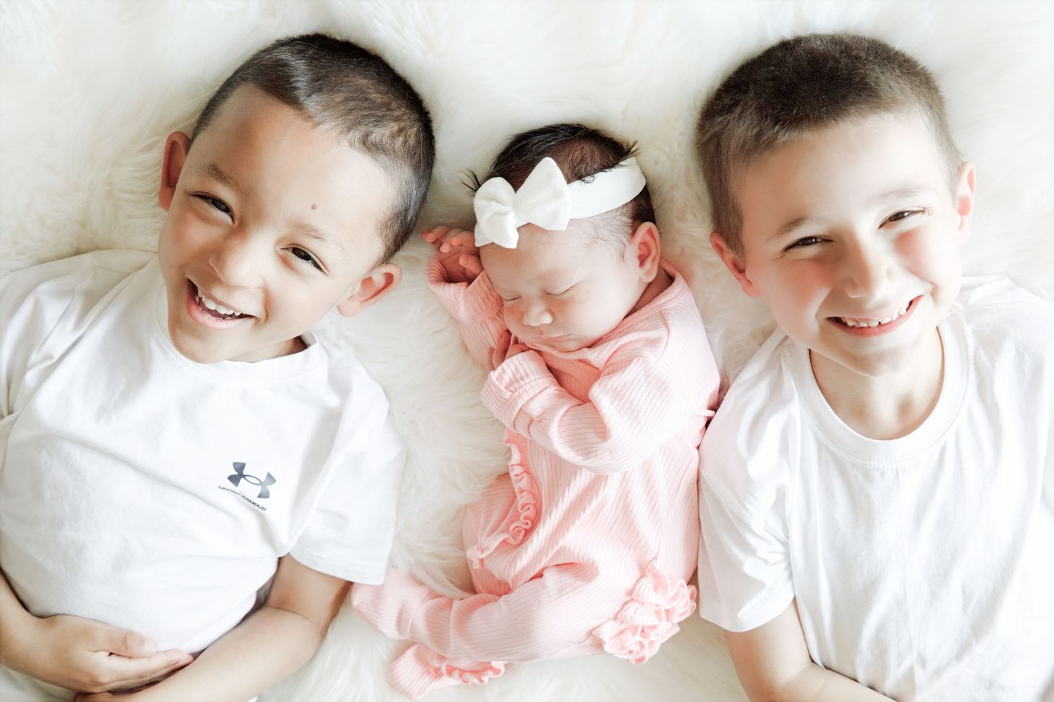 Happy siblings in white shirts posing with baby in pink outfit on white background.