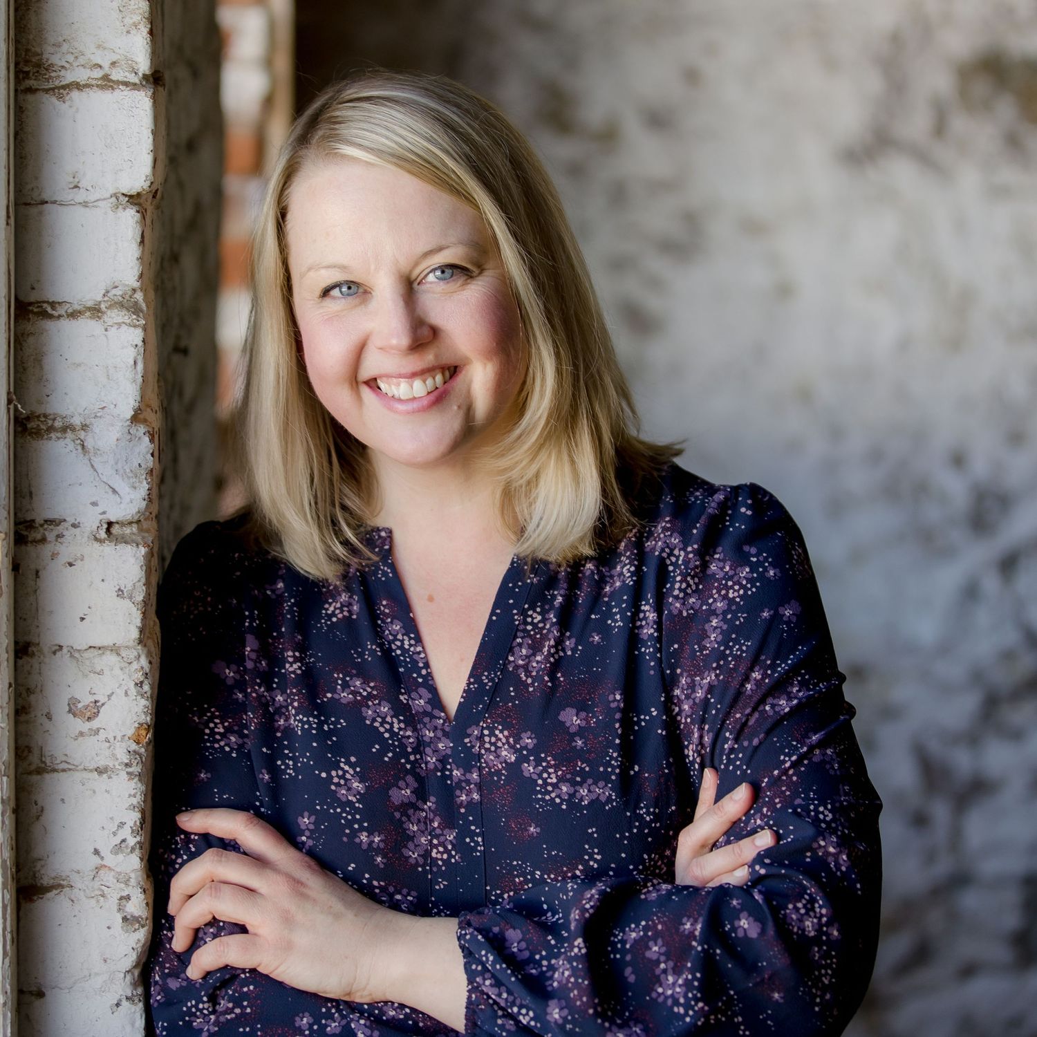 Professional headshot of someone in a navy floral blouse against a textured gray wall background.