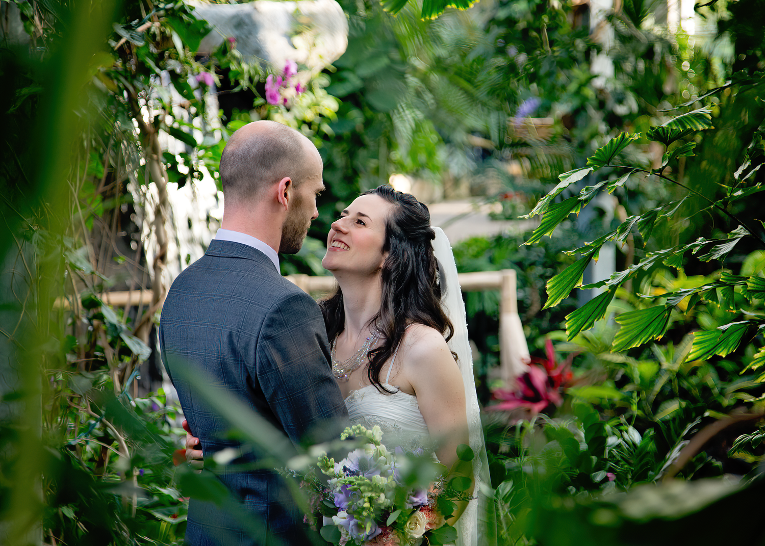 A romantic couple shares an intimate moment in a lush garden setting framed by green foliage and natural light.