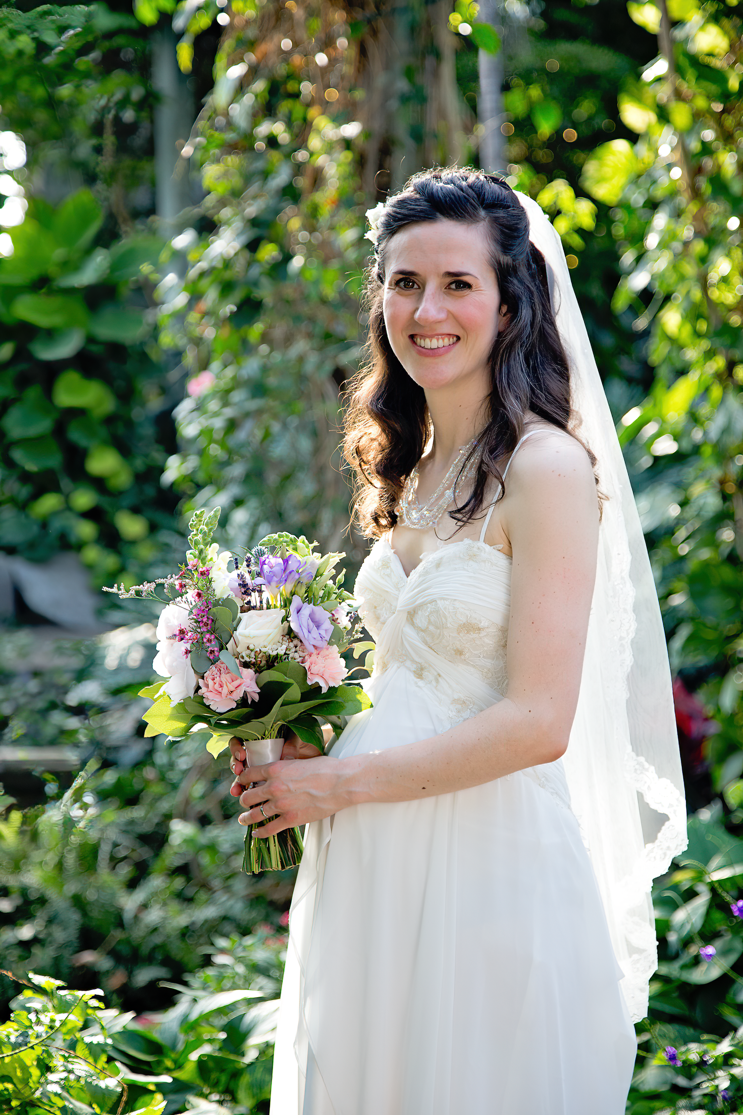 A bride in a flowing white dress holds a colorful bouquet while standing in a lush garden setting with dappled sunlight.