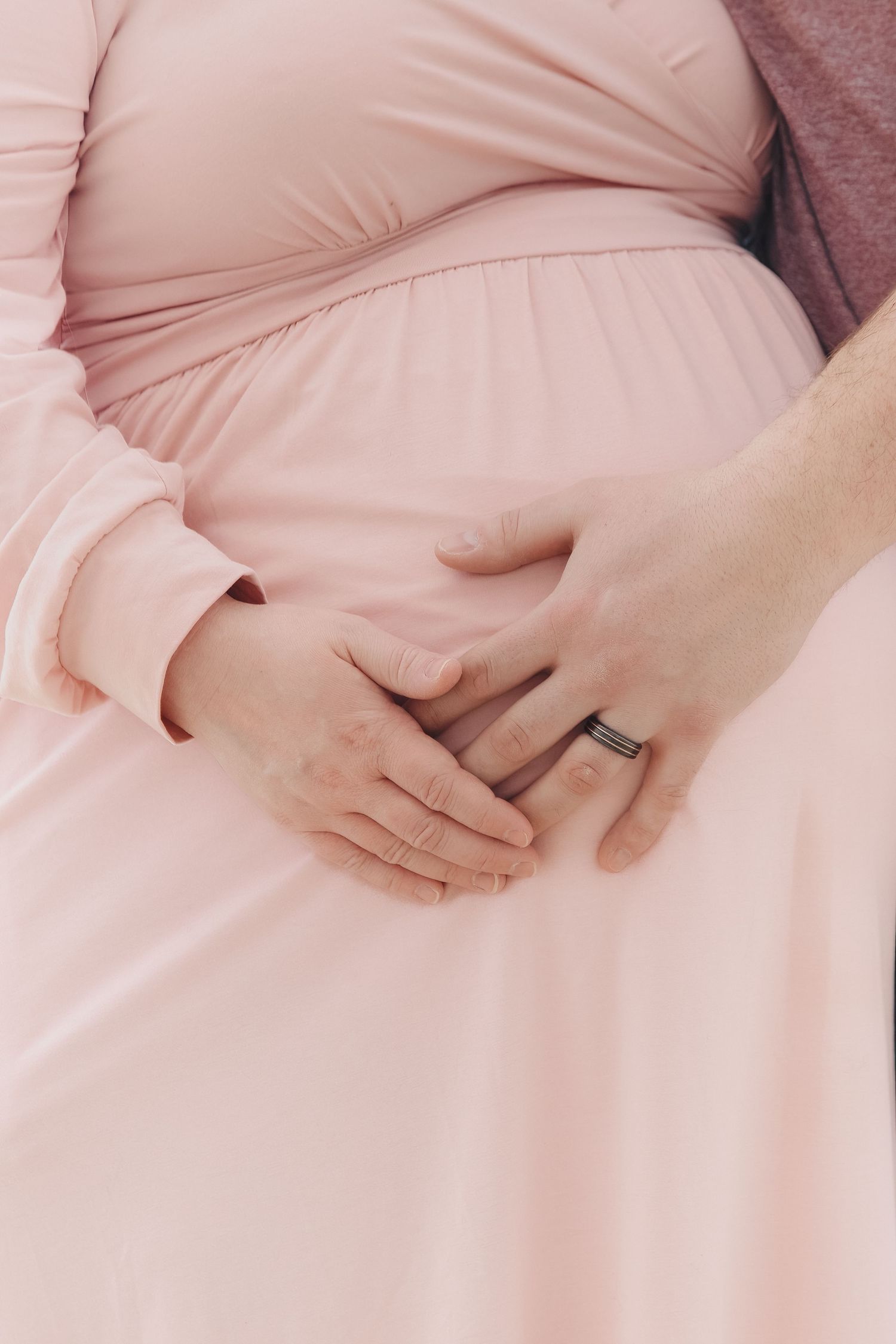 A pregnant woman in a soft pink dress gently cradles her baby bump while wearing a wedding ring.