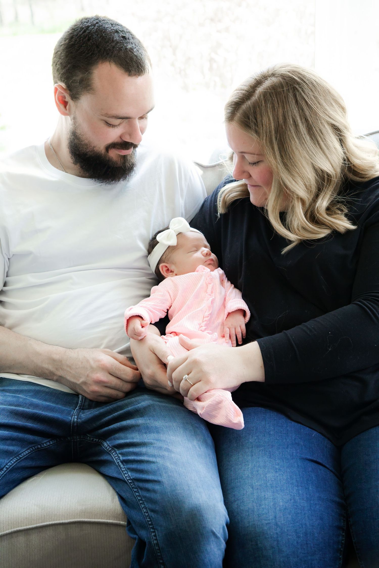 New parents sitting on a couch together, lovingly gazing at their newborn baby girl dressed in pink.