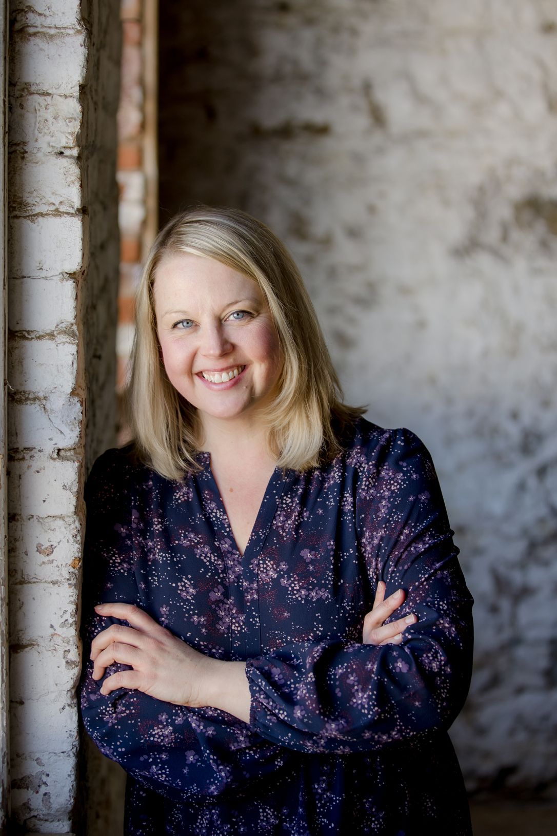 Professional portrait of a woman in a sparkly navy blue top smiling against a rustic concrete wall.