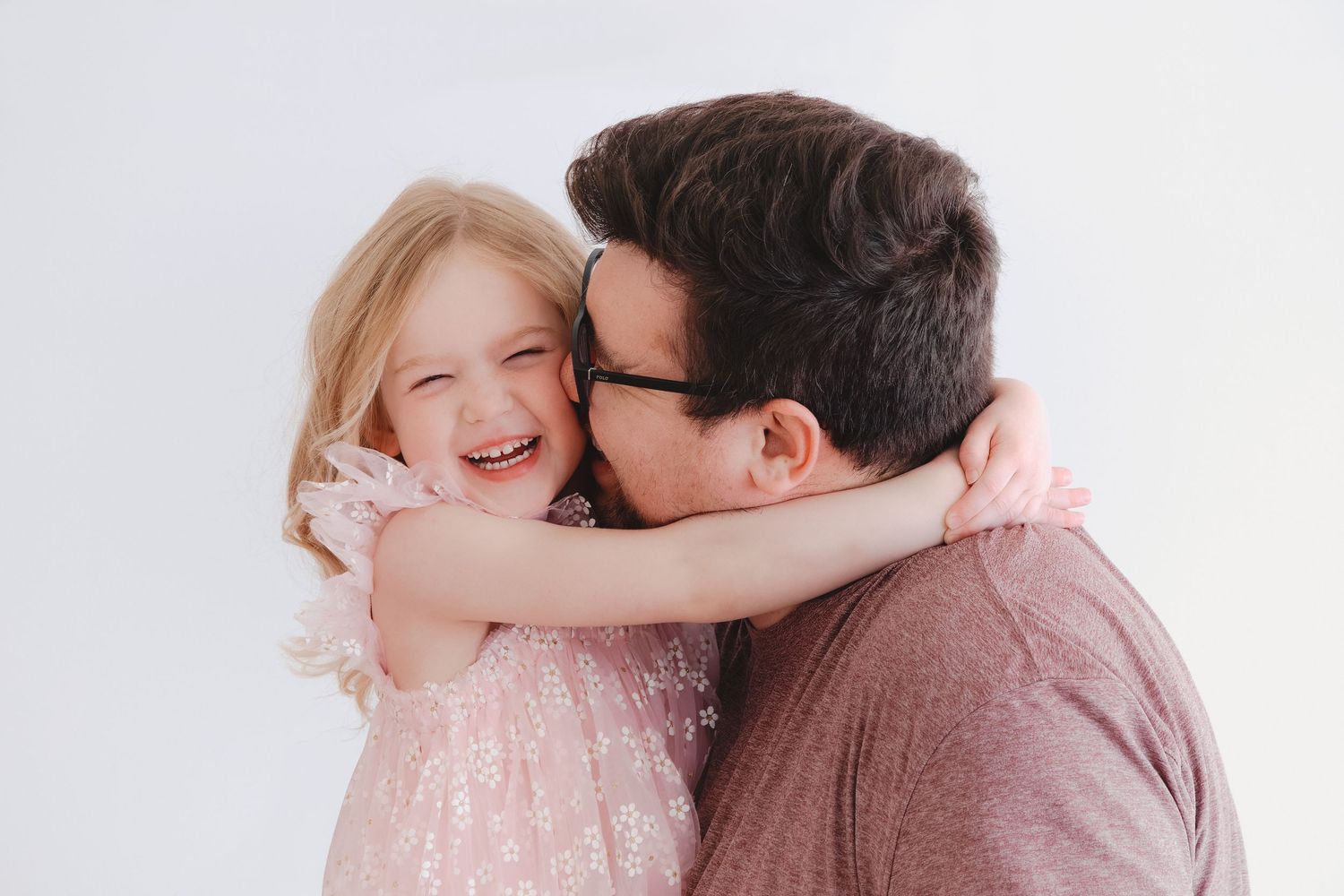 A father kisses his laughing young daughter in a pink dress against a white background.