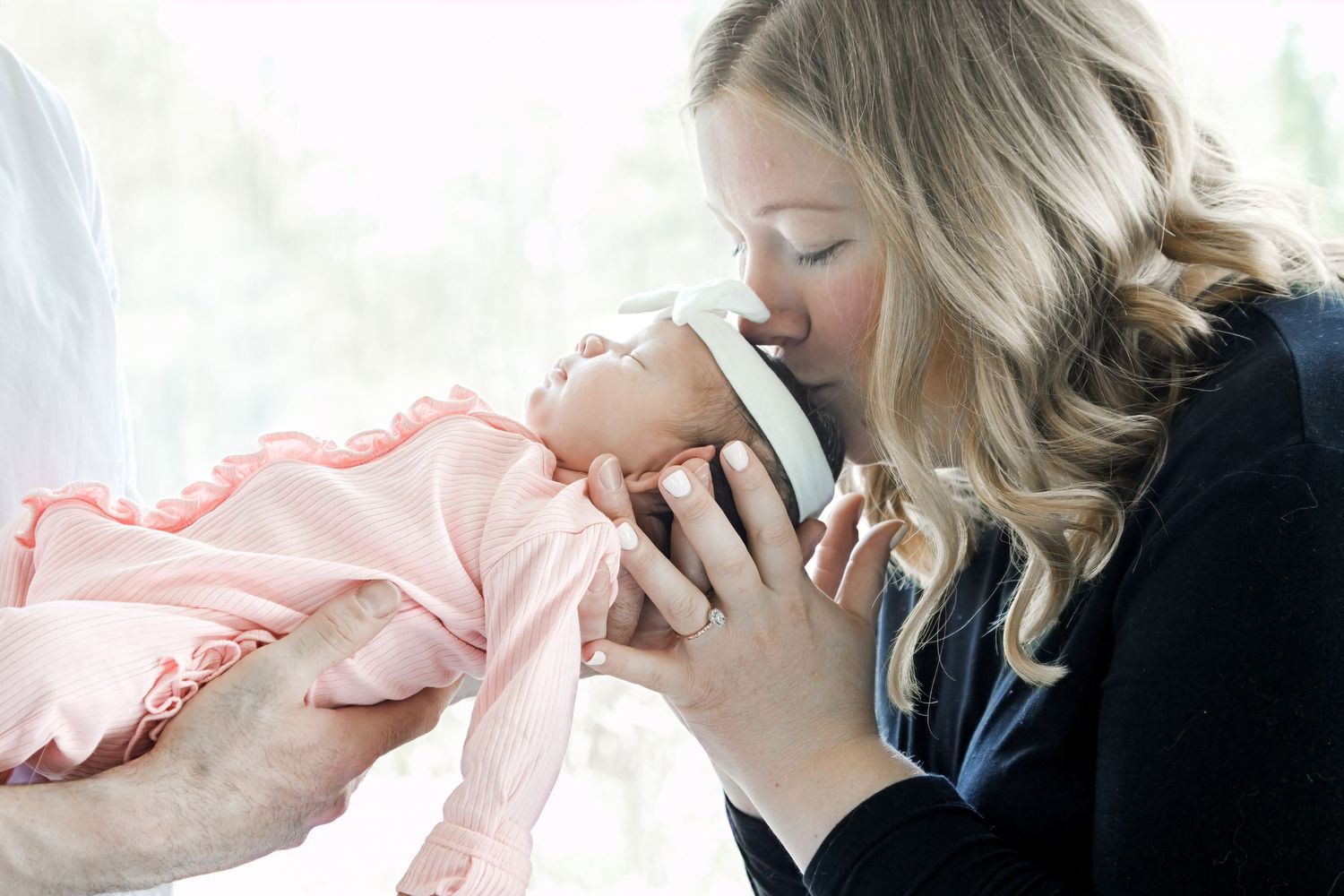 A blonde woman tenderly kisses a newborn baby dressed in pink, cradled in someone's hands.