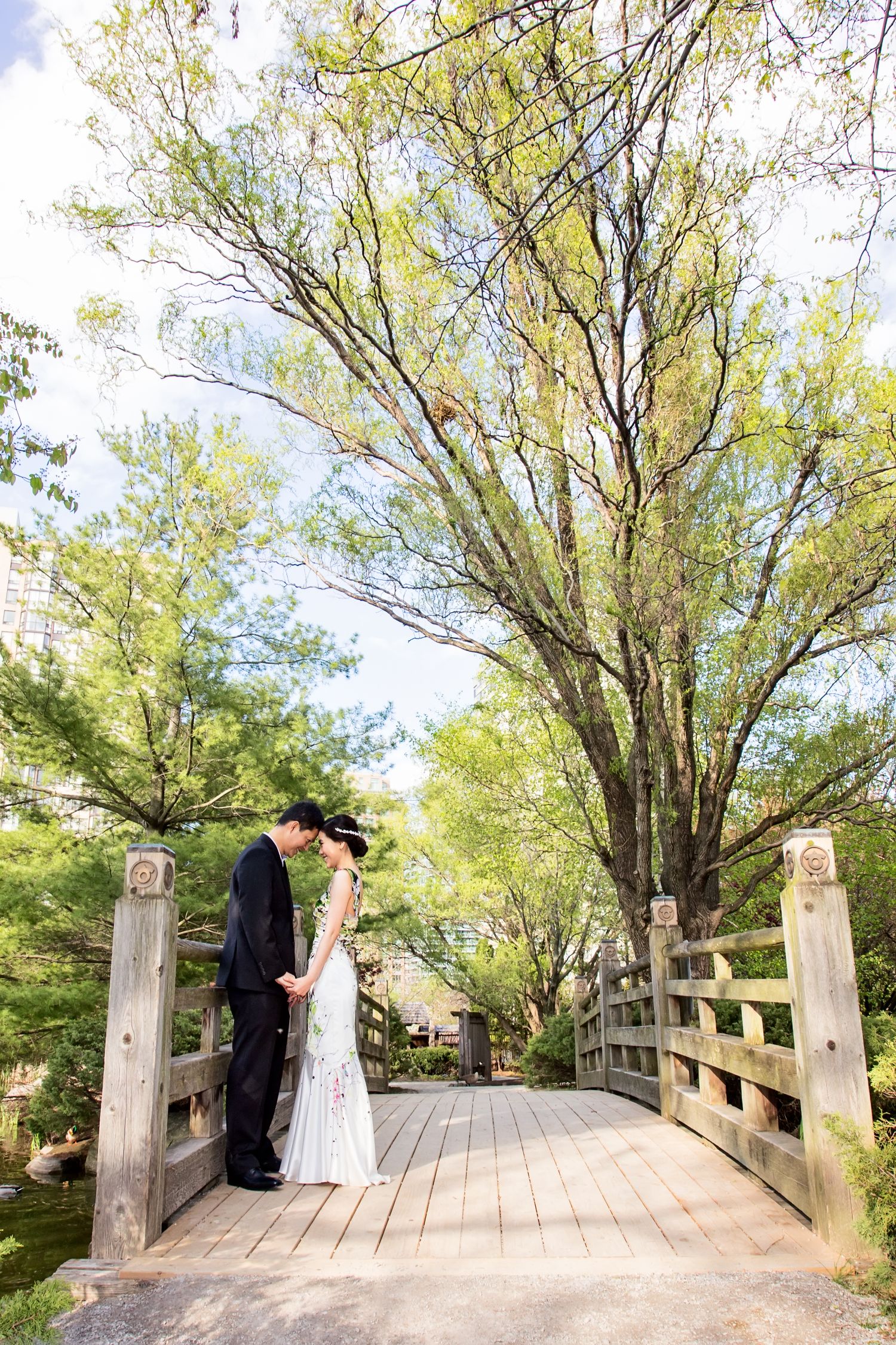 A couple stands on a rustic wooden footbridge surrounded by lush green trees in a scenic outdoor setting.