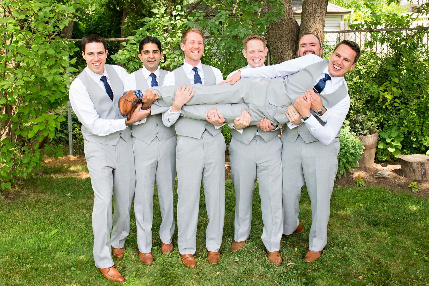 Groomsmen in light gray suits pose playfully in a garden setting for wedding party photos.
