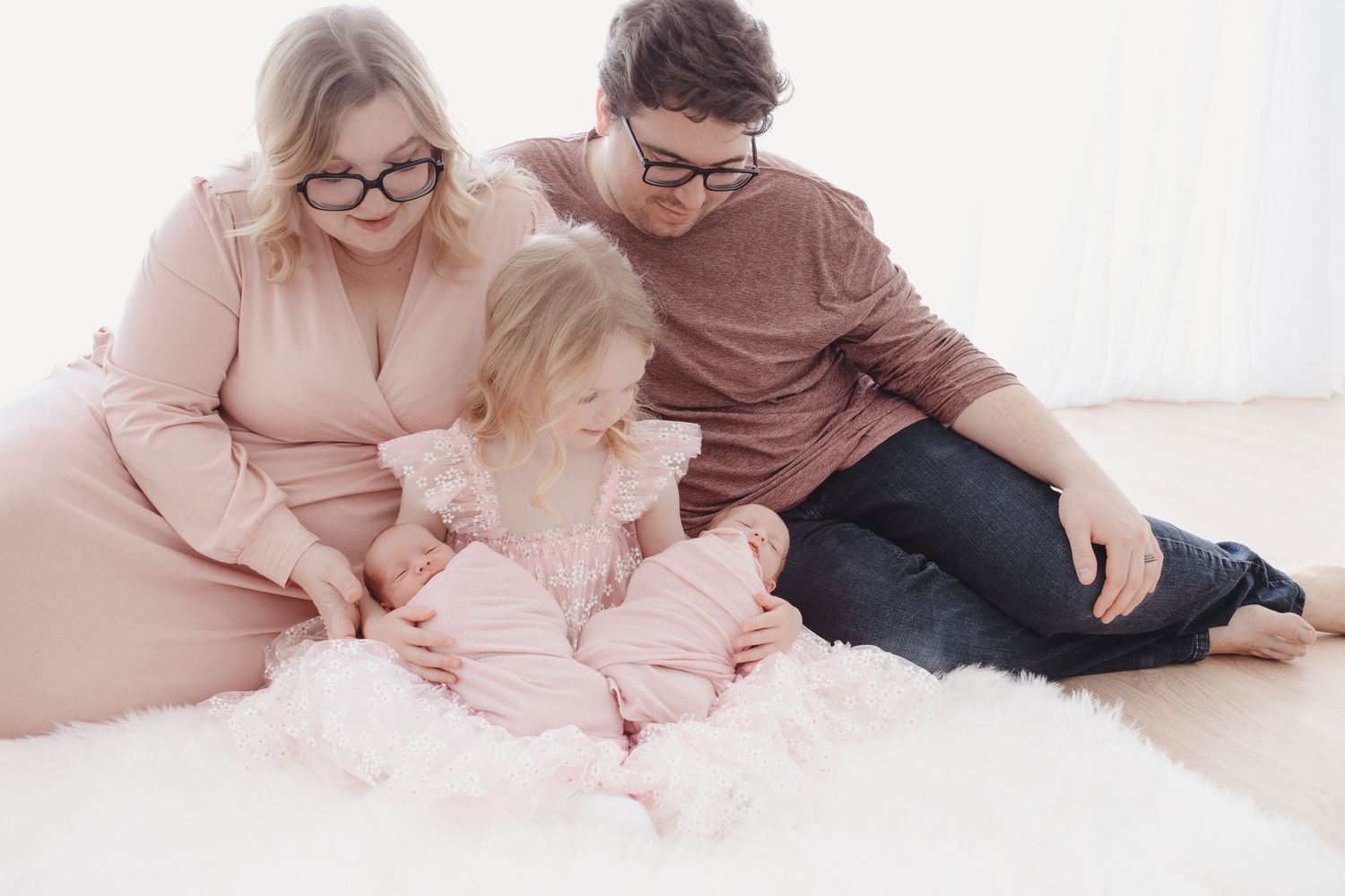 Family with toddler and newborn baby in pink outfits sitting on white fur rug in bright studio.