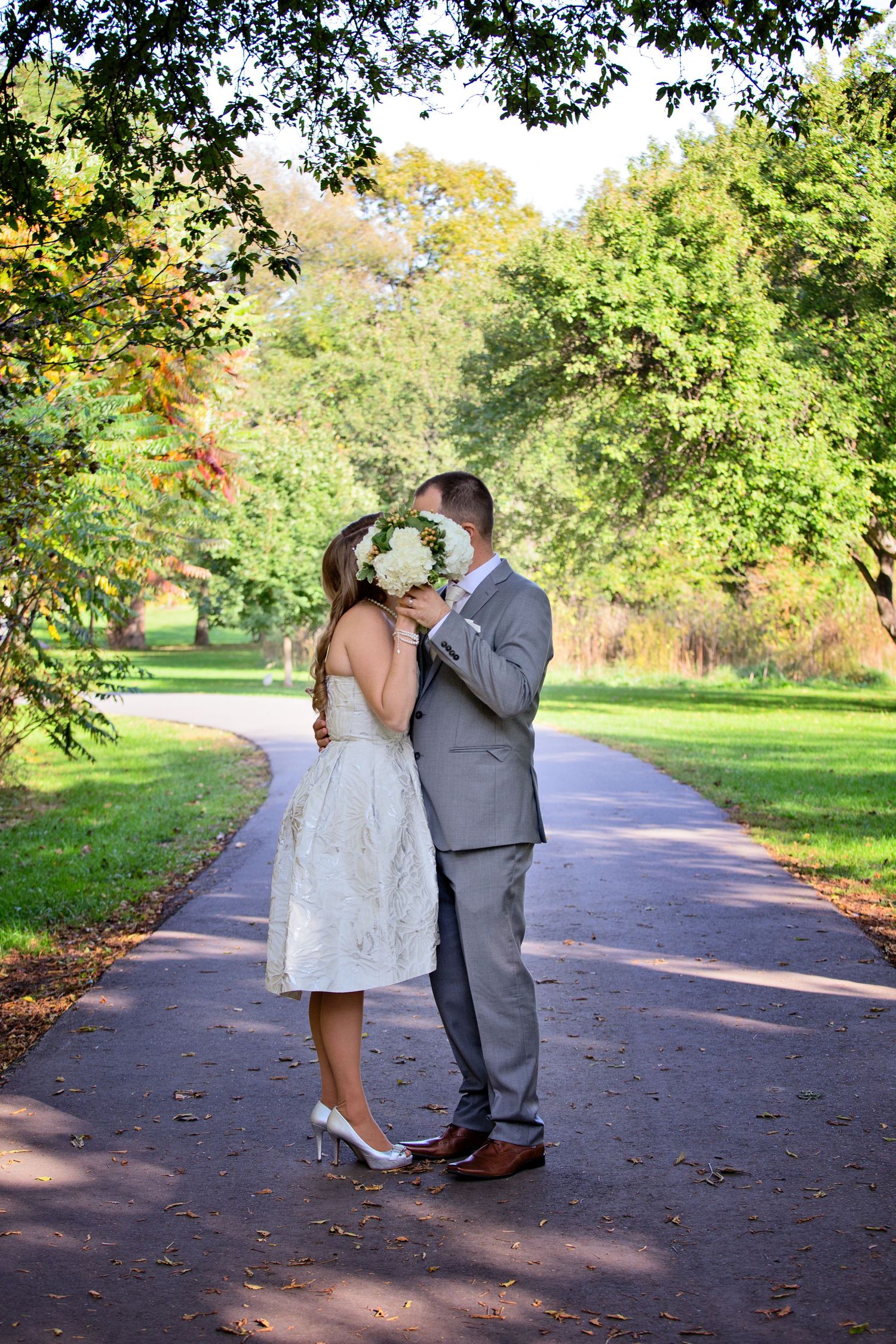 A couple in wedding attire shares a romantic moment on a tree-lined path during an autumn day.