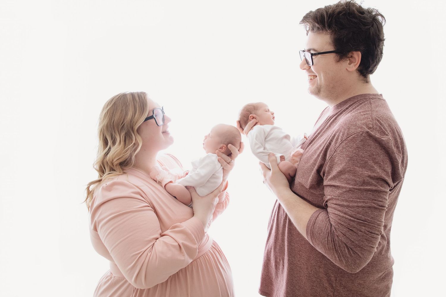 Two parents wearing glasses hold newborn twins facing each other in a bright white studio photo.