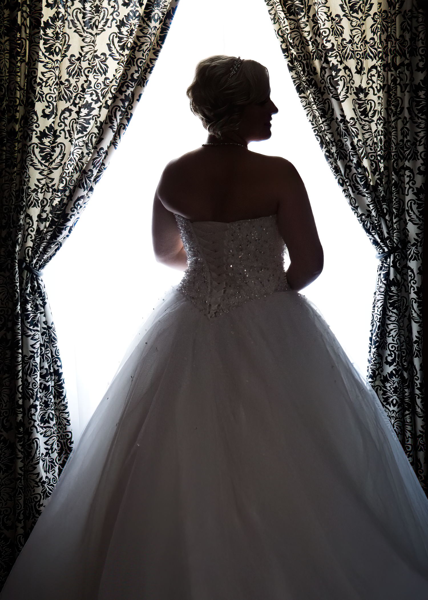 Silhouettes of wedding dresses displayed against ornate lace curtains with dramatic backlighting creating an ethereal effect.