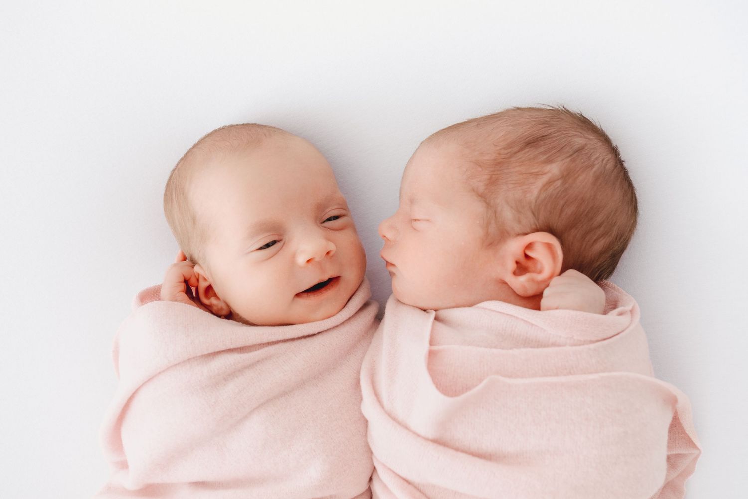 Two newborn twins swaddled in pink blankets lying side by side on a white background, one smiling.