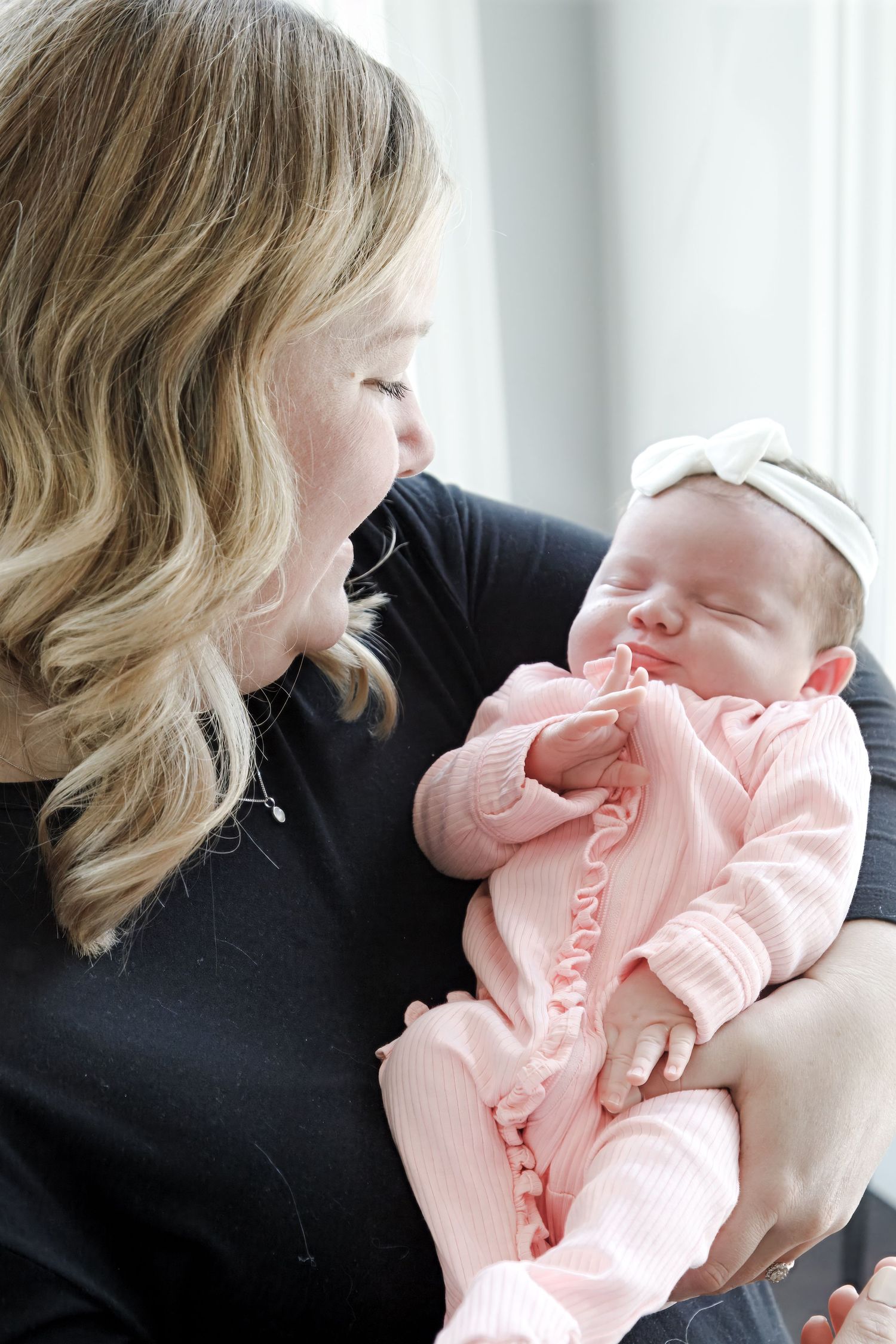 A blonde woman in a black top lovingly gazes at her sleeping newborn baby dressed in a pink ruffled outfit.