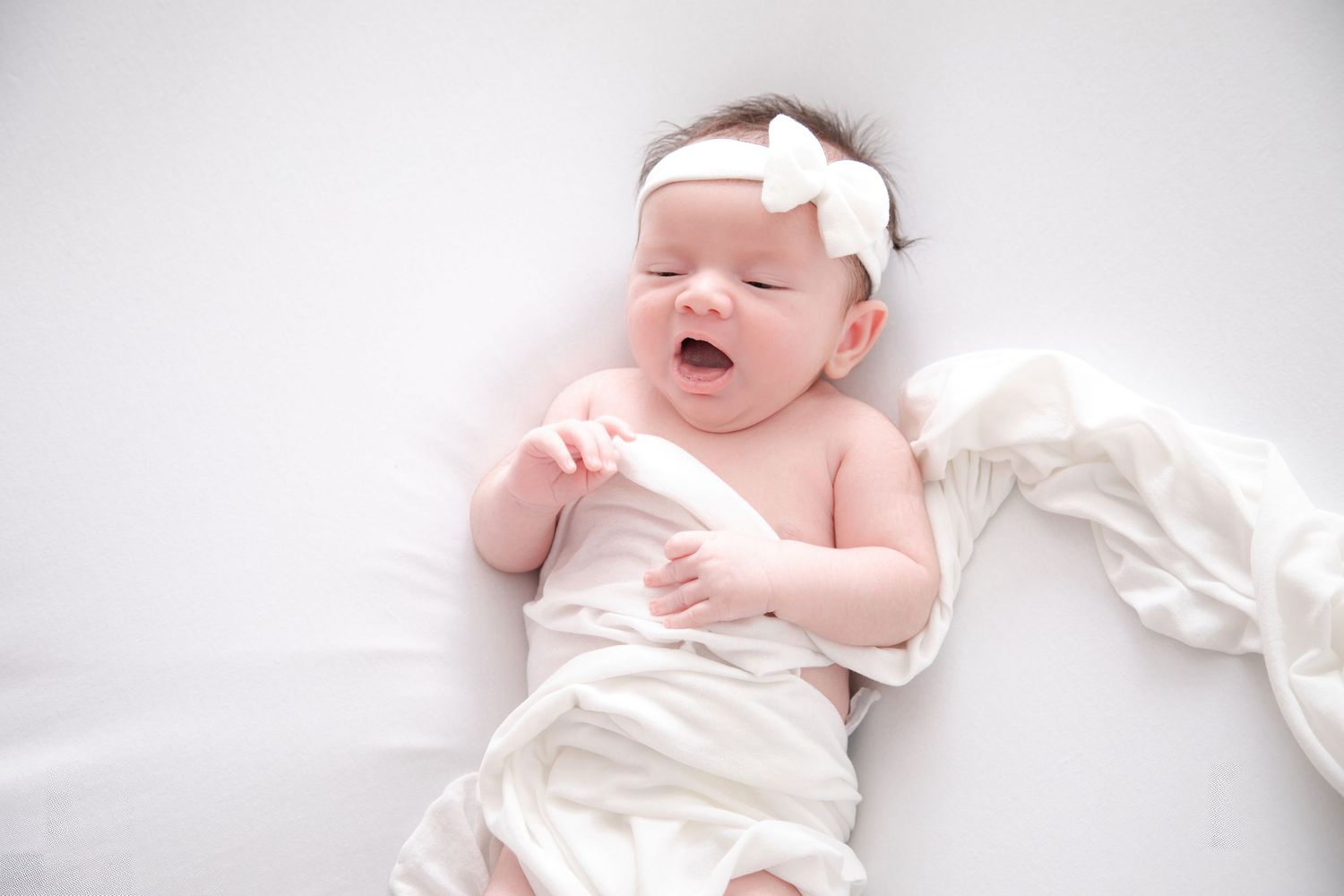 Newborn wearing white bow headband crying on light background.