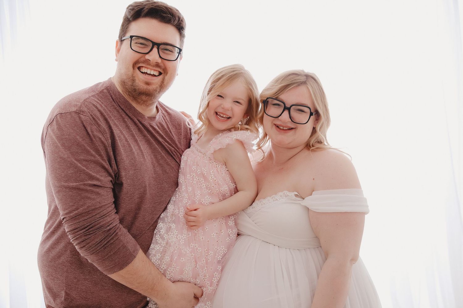 Happy family of three posing for maternity photo, parents wearing glasses, young daughter in pink dress.