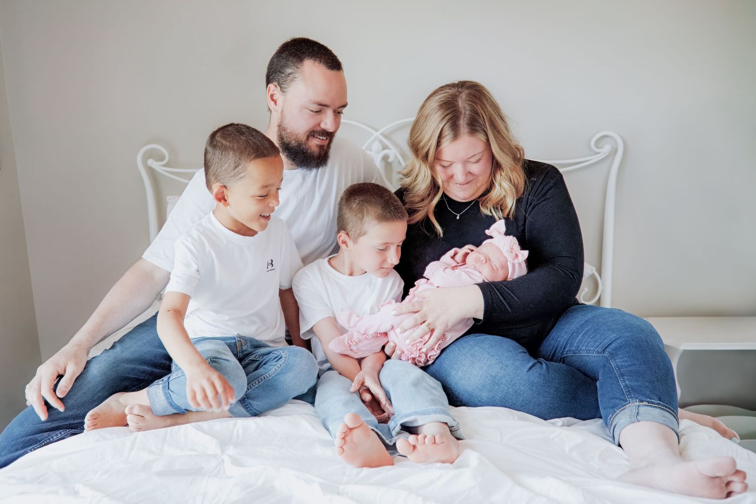 Happy family of five sitting on a bed, parents with two young boys and a newborn baby girl in pink.