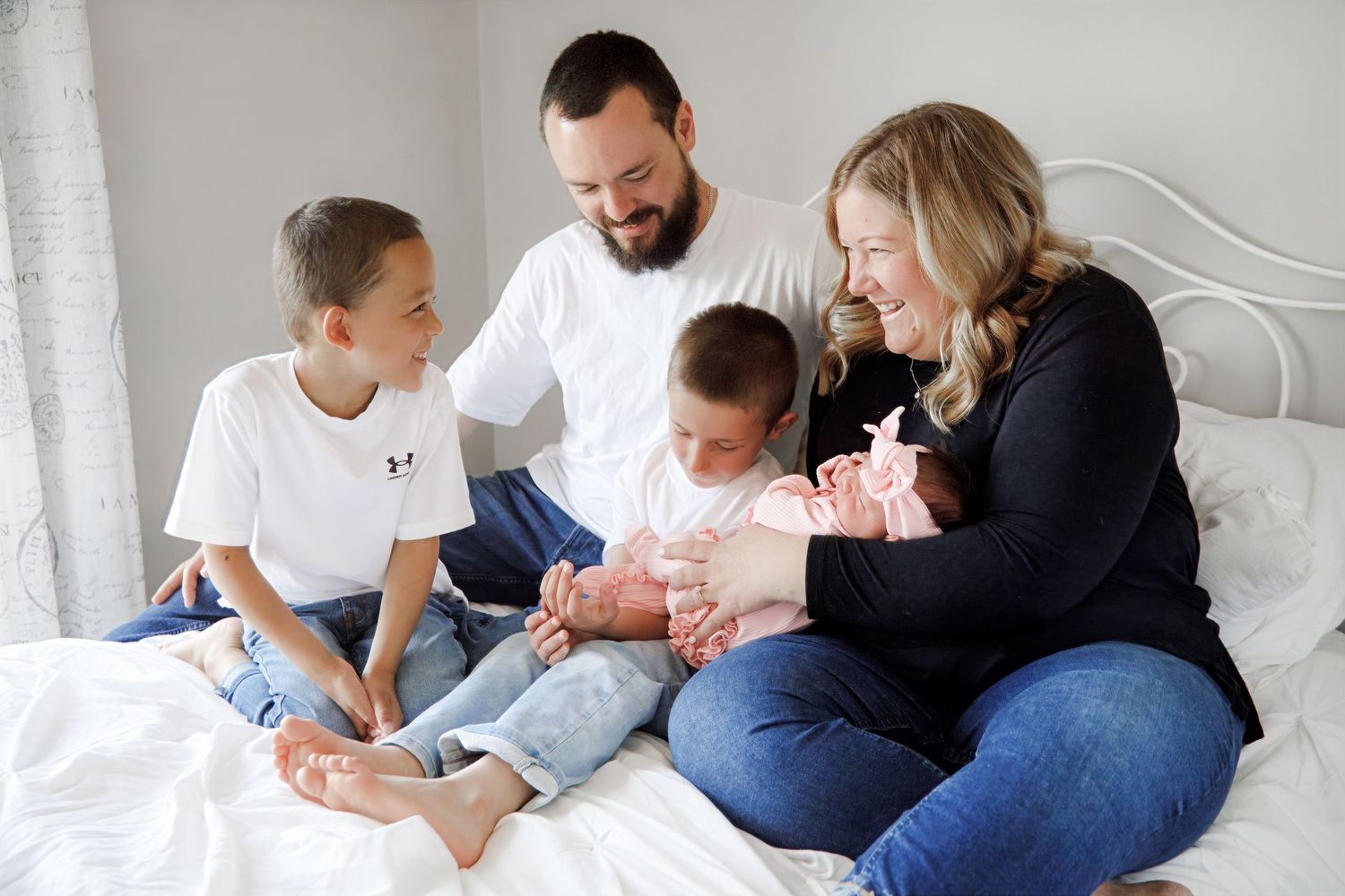 Happy family of five sitting on a white bed, parents smiling at newborn baby girl in pink, with two young boys nearby.