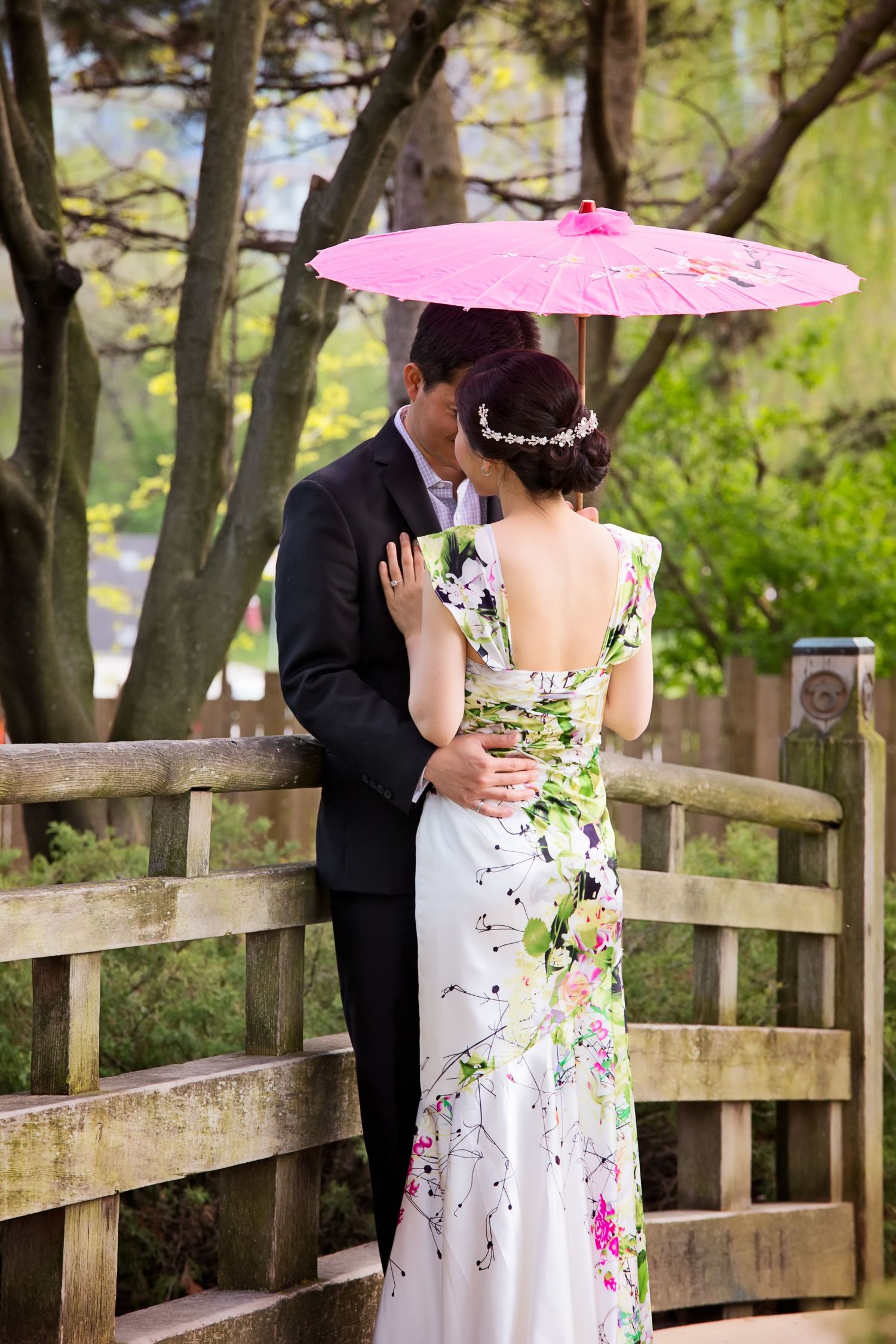 A couple shares a romantic moment on a wooden bridge in a garden setting with a pink parasol.