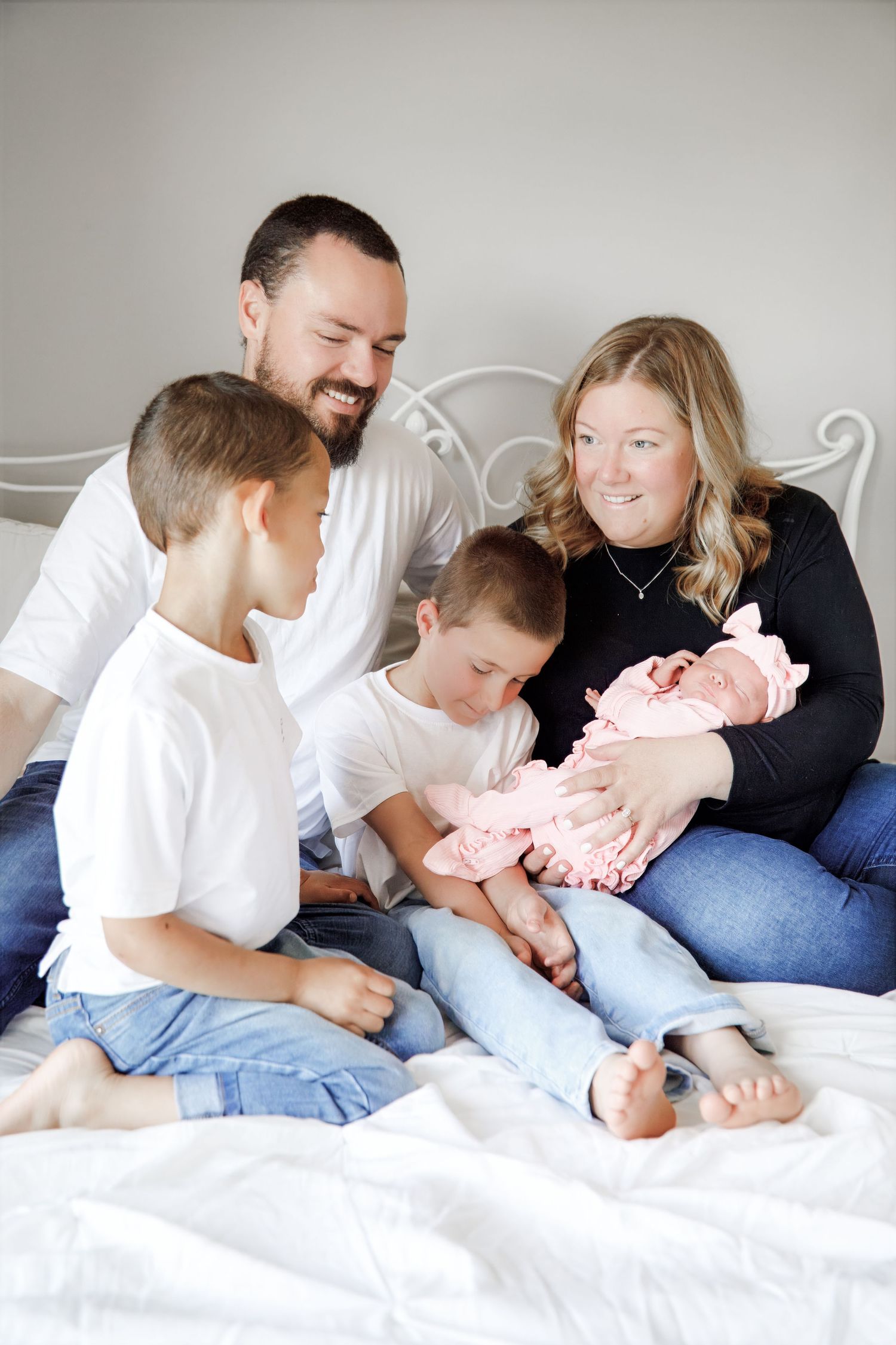 Happy family of five sitting on a bed, parents smiling at newborn baby girl in pink, with two young boys.