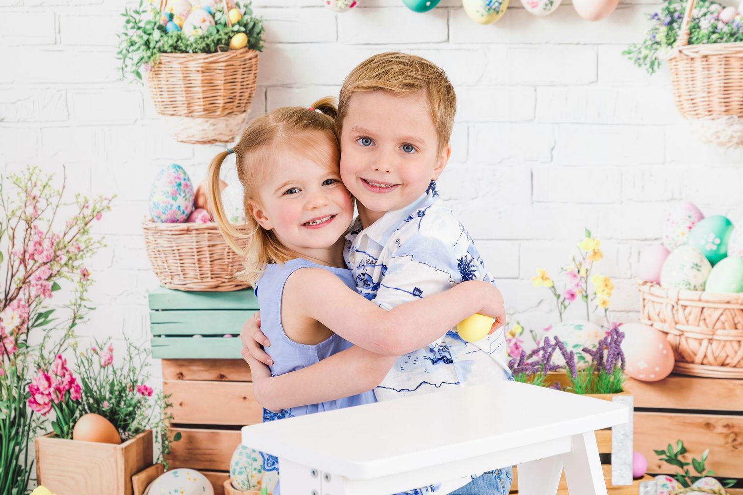 Children in blue and white outfits pose together in a spring-themed studio setting with floral decorations.