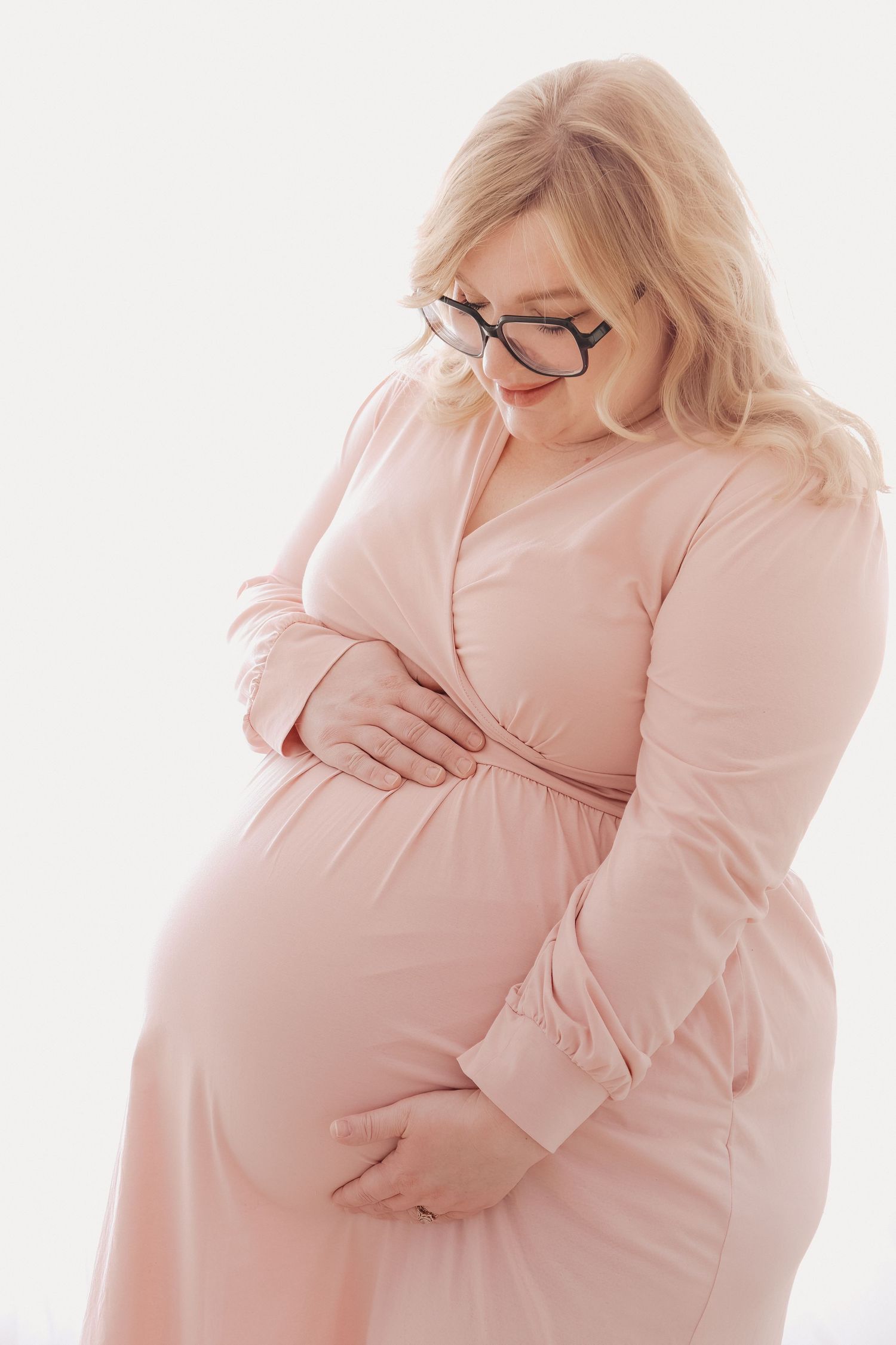 Pregnant woman in pink dress and glasses cradling her baby bump against a white background.
