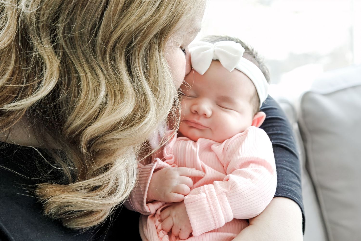 Mother tenderly kissing her sleeping newborn baby girl wearing a pink outfit and white bow headband.