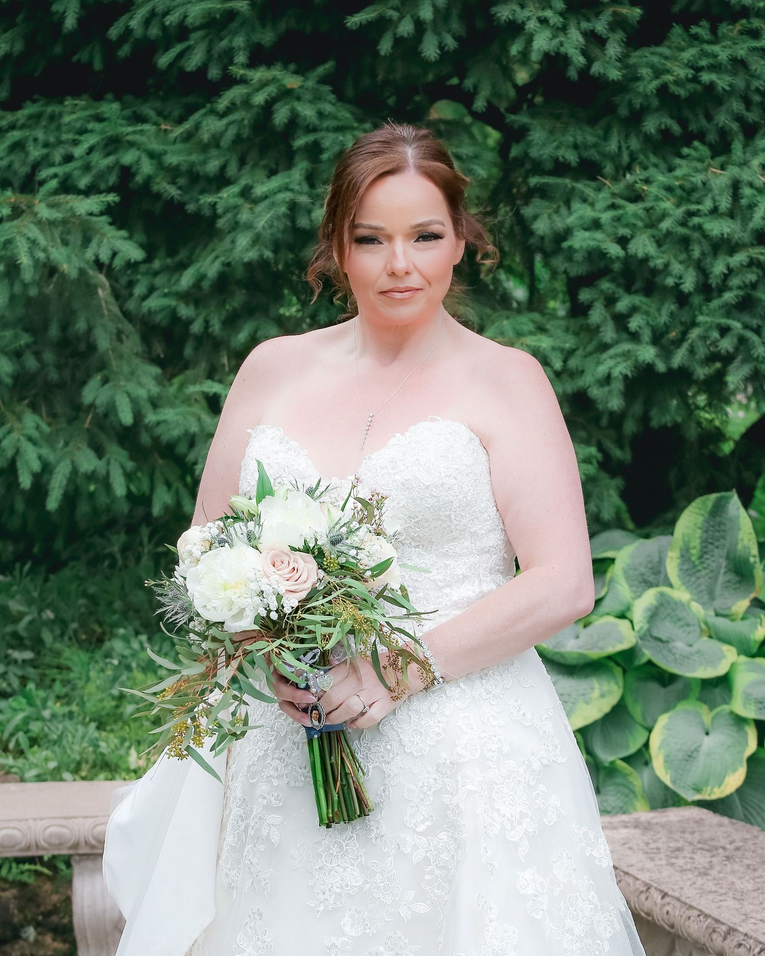 A bride in a white lace wedding dress holds a lush bouquet of white flowers and greenery in an outdoor garden setting.