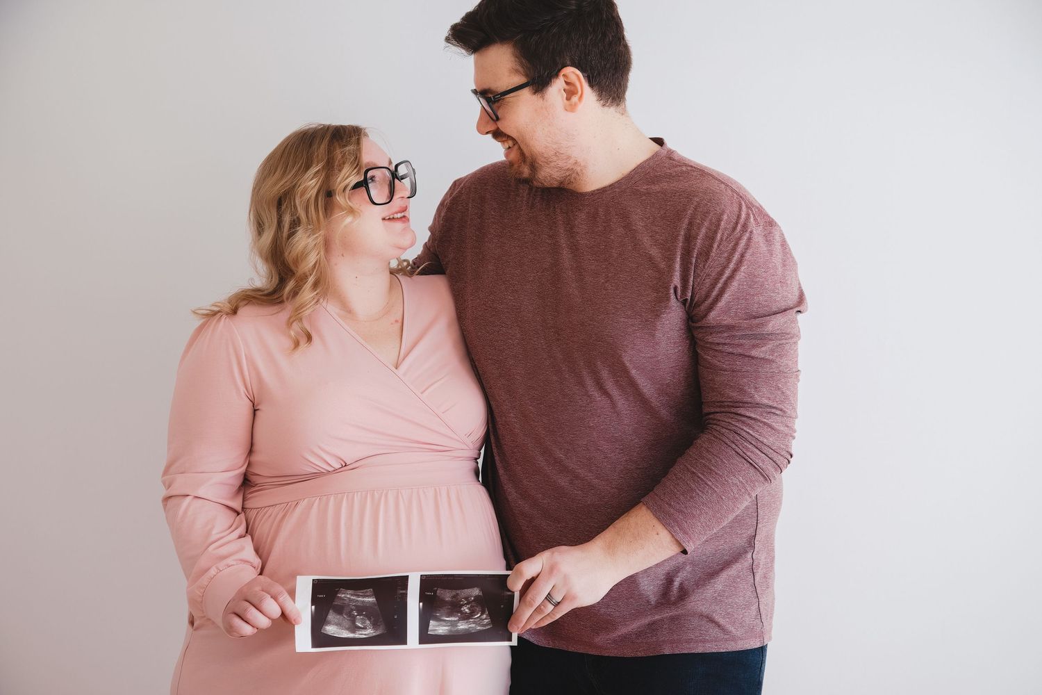 Pregnant couple smiling at each other while holding ultrasound photos against a white background.