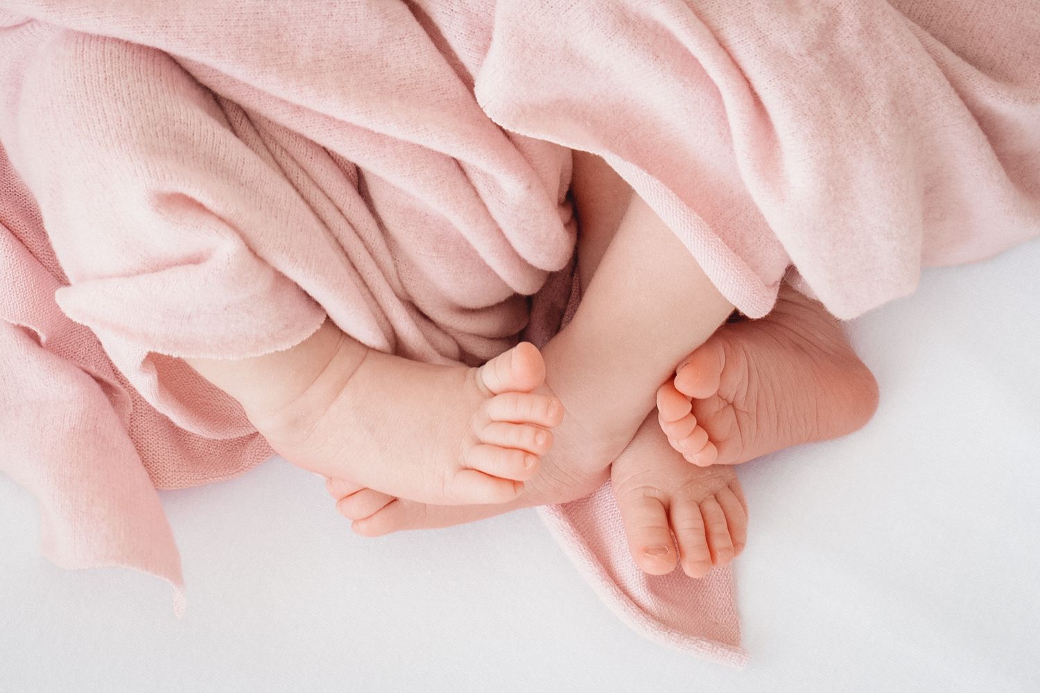 Tiny newborn baby feet peeking out from a soft pink blanket on a white surface.