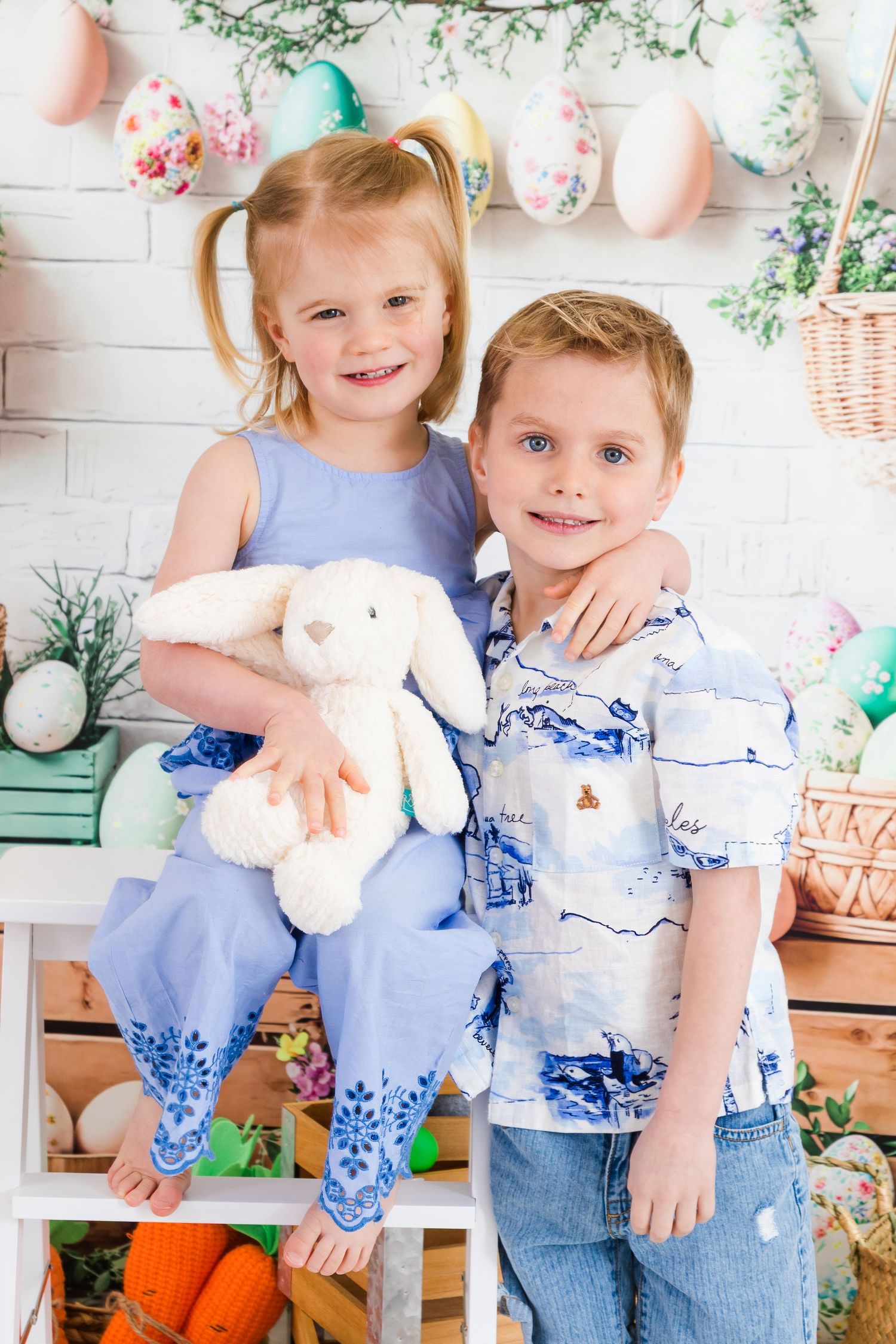 Two young children pose together for an Easter photo with spring decorations, pastel balloons, and a white plush bunny.