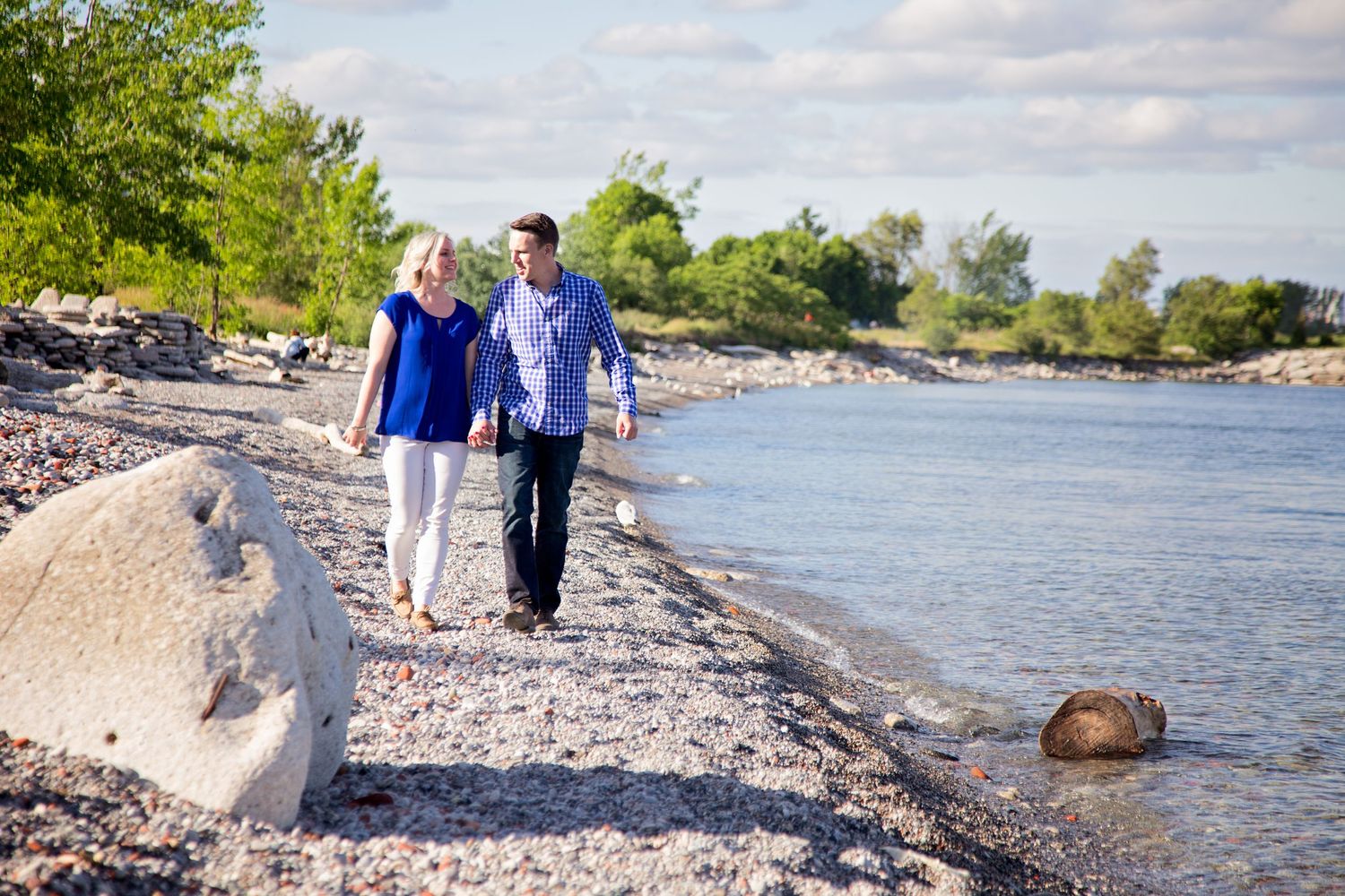 People walking along a rocky shoreline with blue water and trees visible on a sunny summer day.