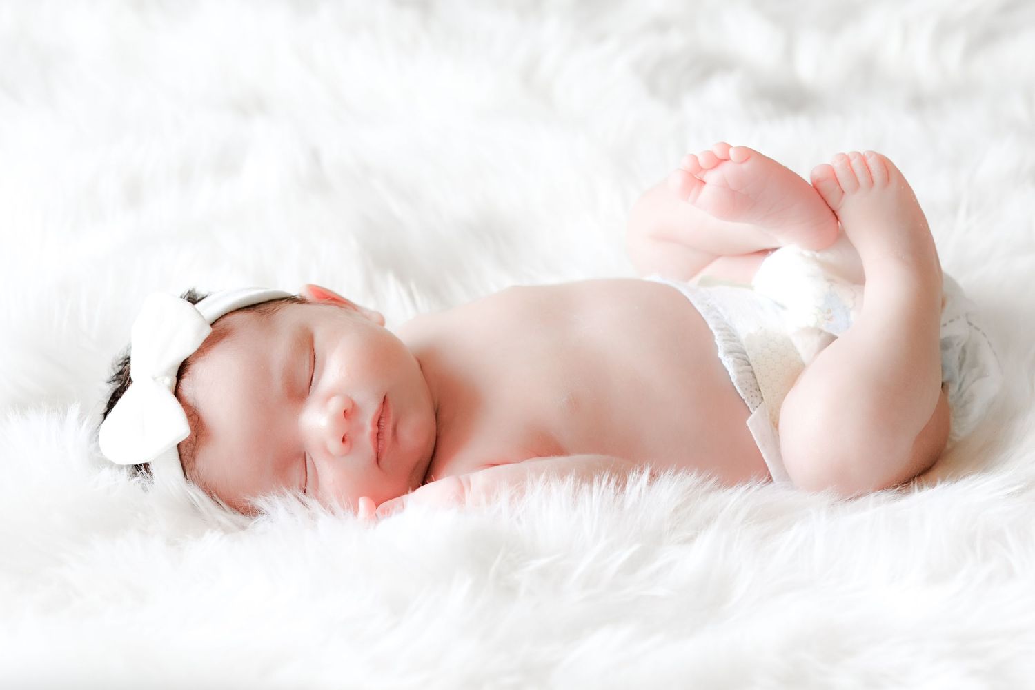 A newborn baby sleeping peacefully on soft white blankets in a bright studio setting.