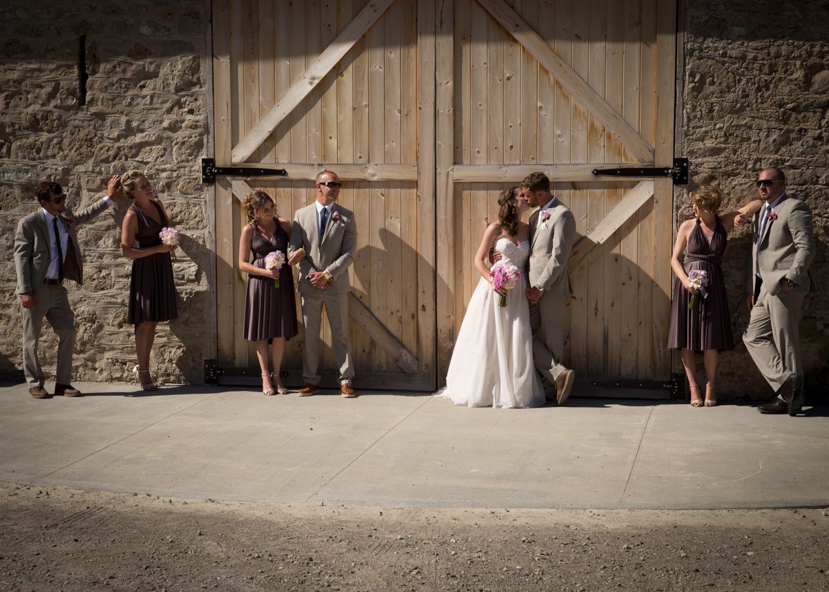 A wedding party stands in front of rustic wooden barn doors for a series of candid photo poses.