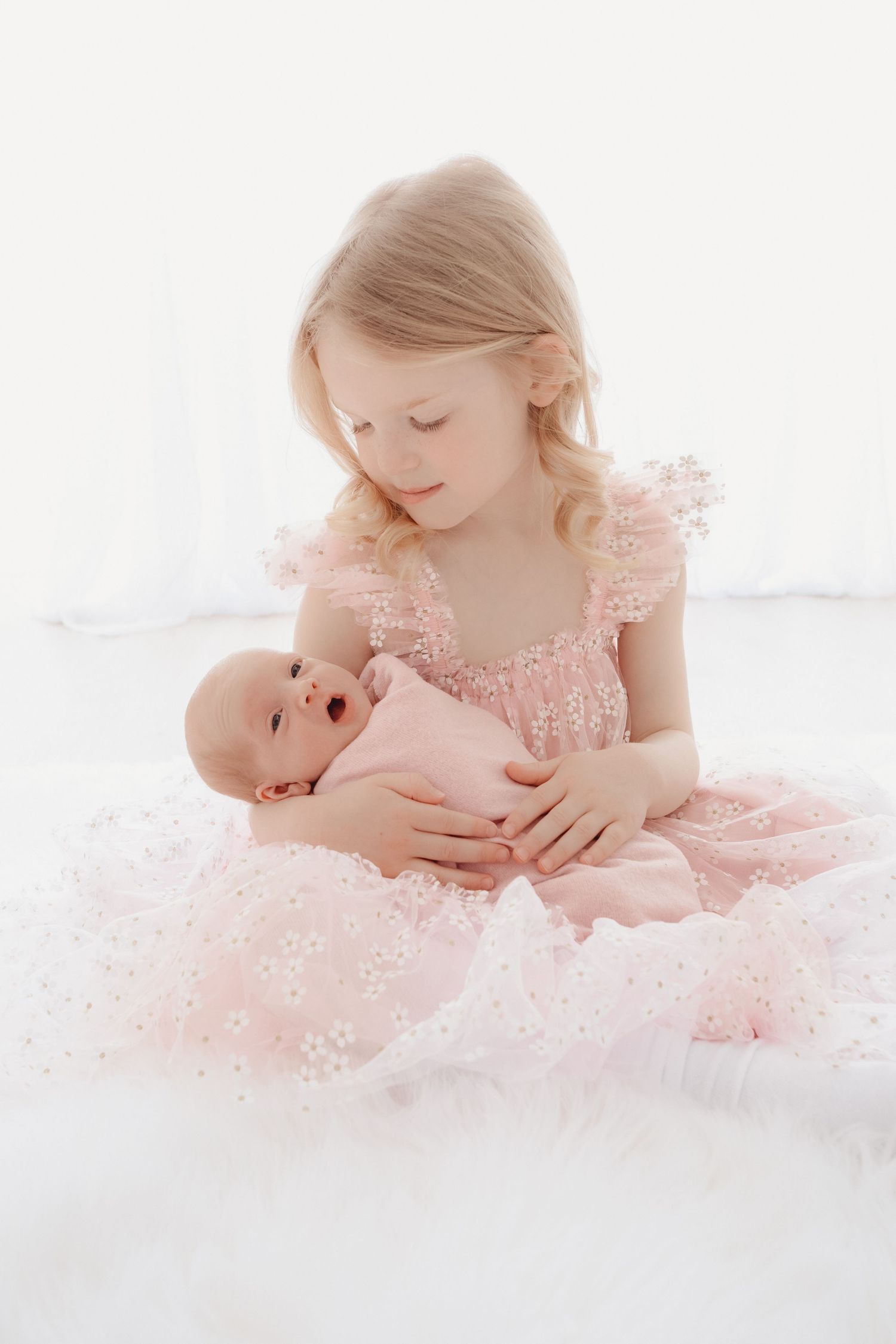 Young girl in pink floral dress gently holds newborn baby sibling on white fur blanket in bright studio.
