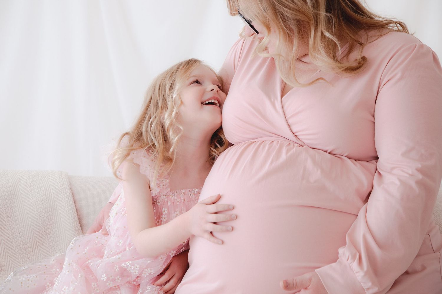 A pregnant woman in pink dress with her young daughter in matching pink dress touching her baby bump.