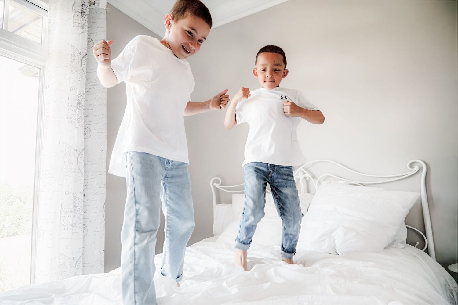 Two people in white shirts and jeans jumping playfully on a white bed near a bright window.