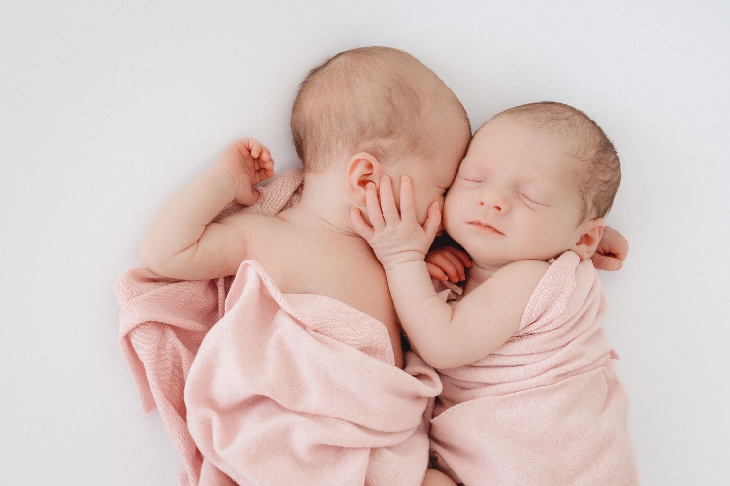Two newborn twins sleeping peacefully together wrapped in soft pink blankets on a white background.