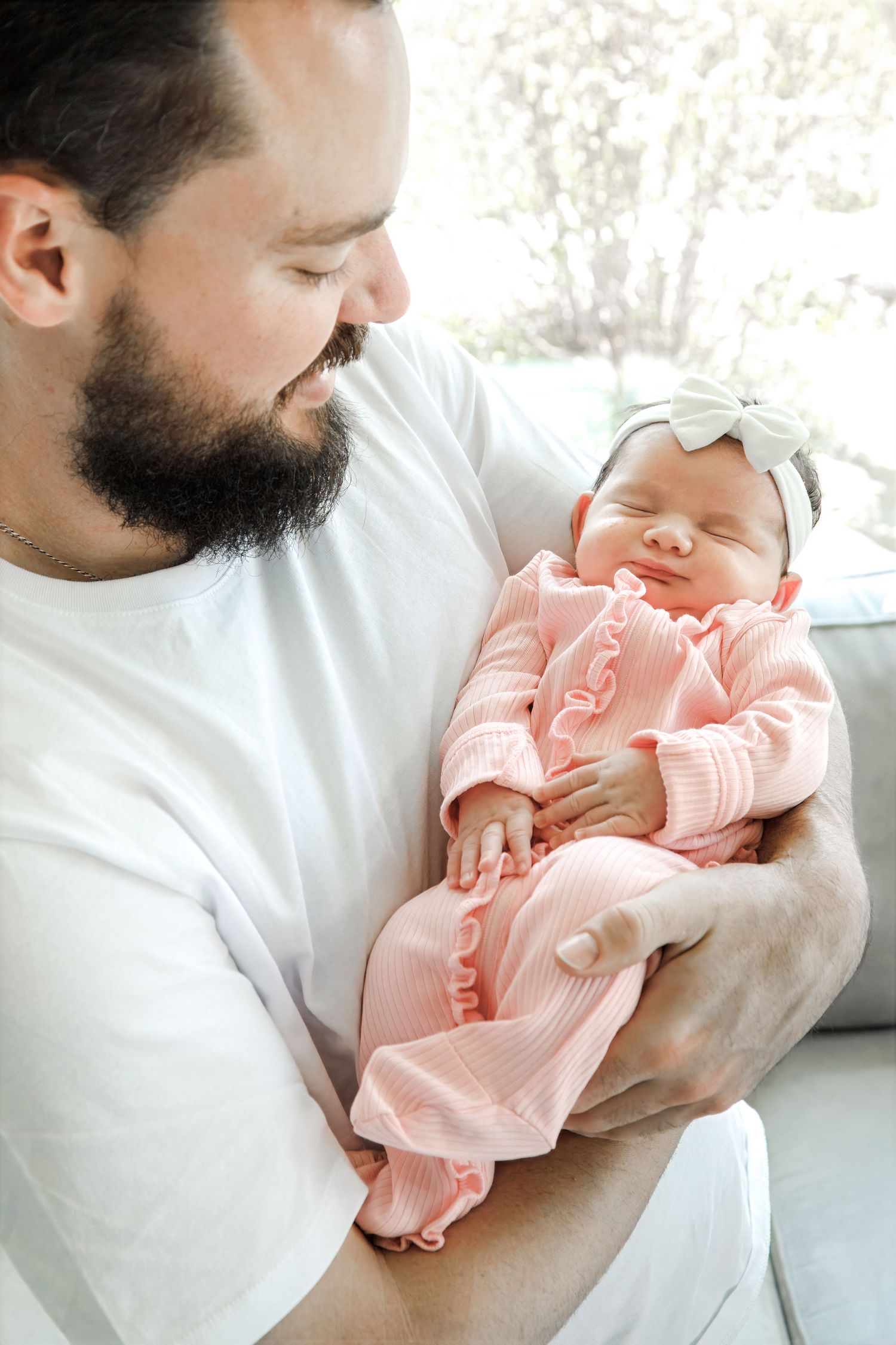 Bearded father in white t-shirt tenderly holds sleeping newborn baby girl in pink ruffle outfit with white bow headband.