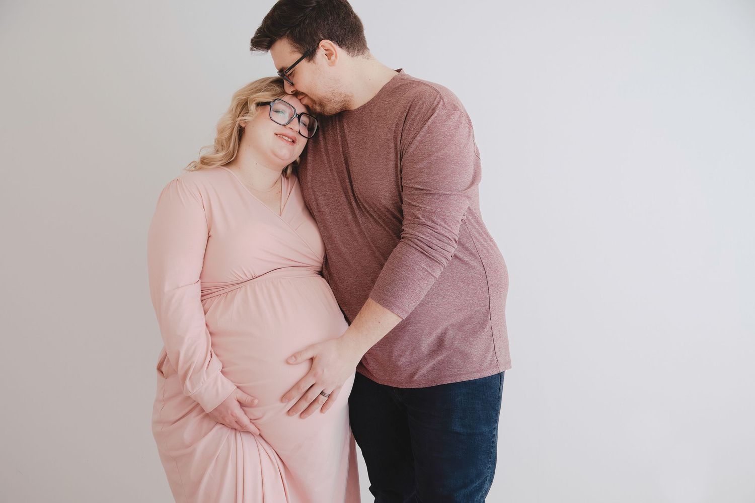 Pregnant couple posing together, woman in pink dress cradling baby bump, man in mauve shirt leaning in tenderly.