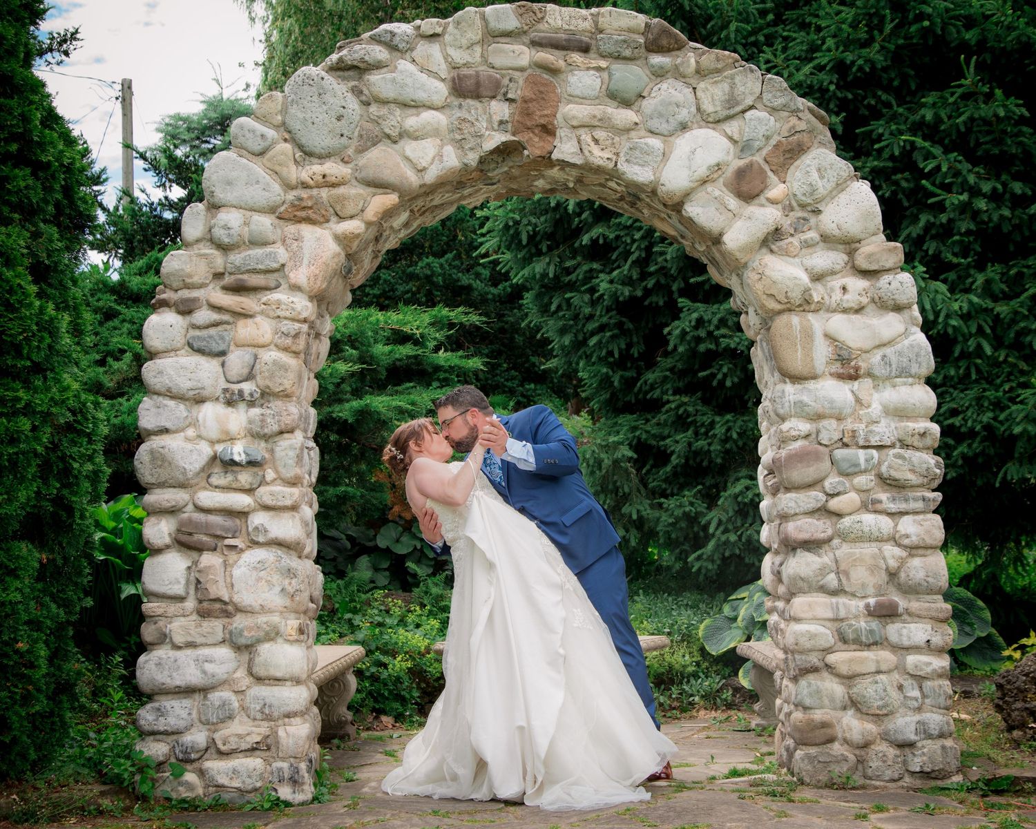 A romantic couple shares a tender moment under a rustic stone archway in a lush garden setting.