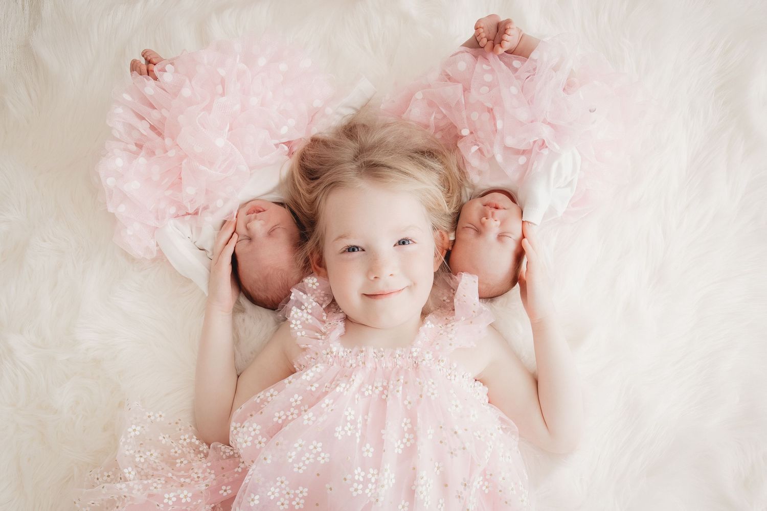 Young girl in pink dress smiles while lying between two newborn twins on a fluffy white blanket.