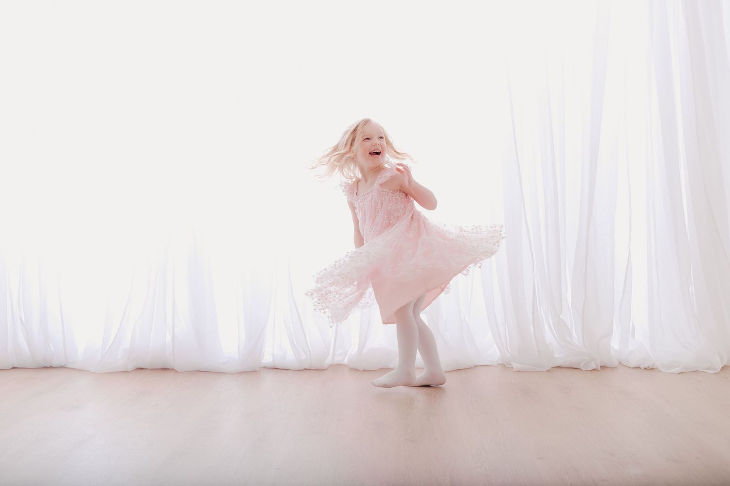 A joyful young girl in a pink tutu dress twirls and dances in a bright white room with wooden floors.