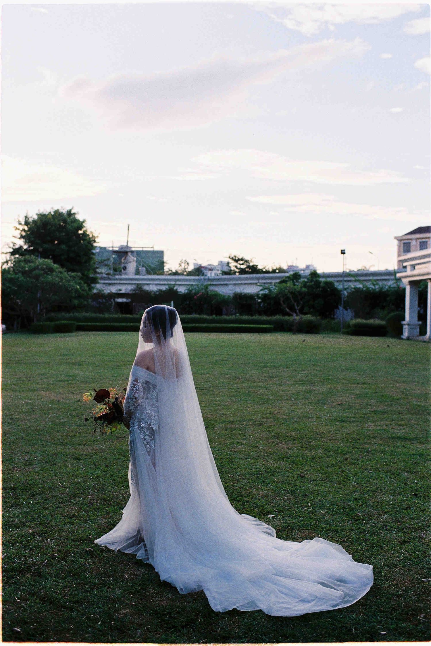 Bride in a lace gown with long veil holds bouquet on green lawn at dusk, back to camera.