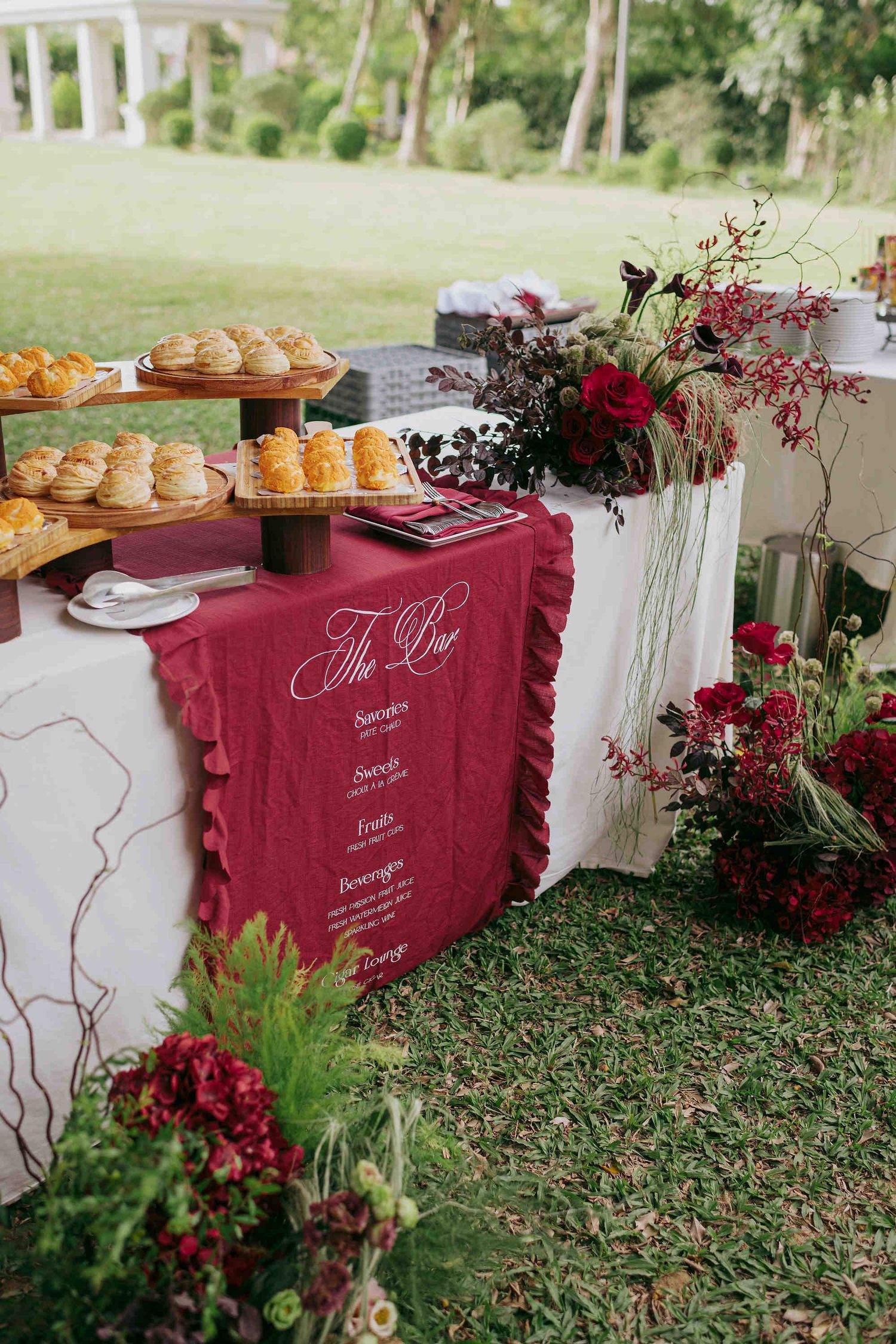 Elegant outdoor wedding buffet table with crimson ruffled runner, floral arrangements, and tiered pastry display on green lawn.
