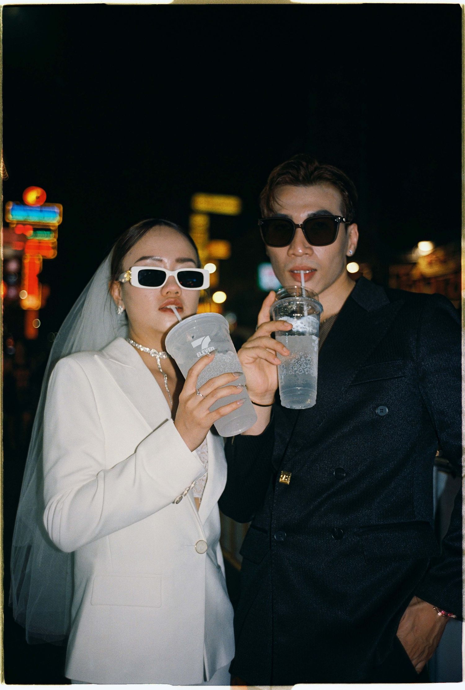 Saigon pre-wedding: two people in contrasting outfits share drinks on a nighttime street with neon signs in background, by AnhVuWedding.