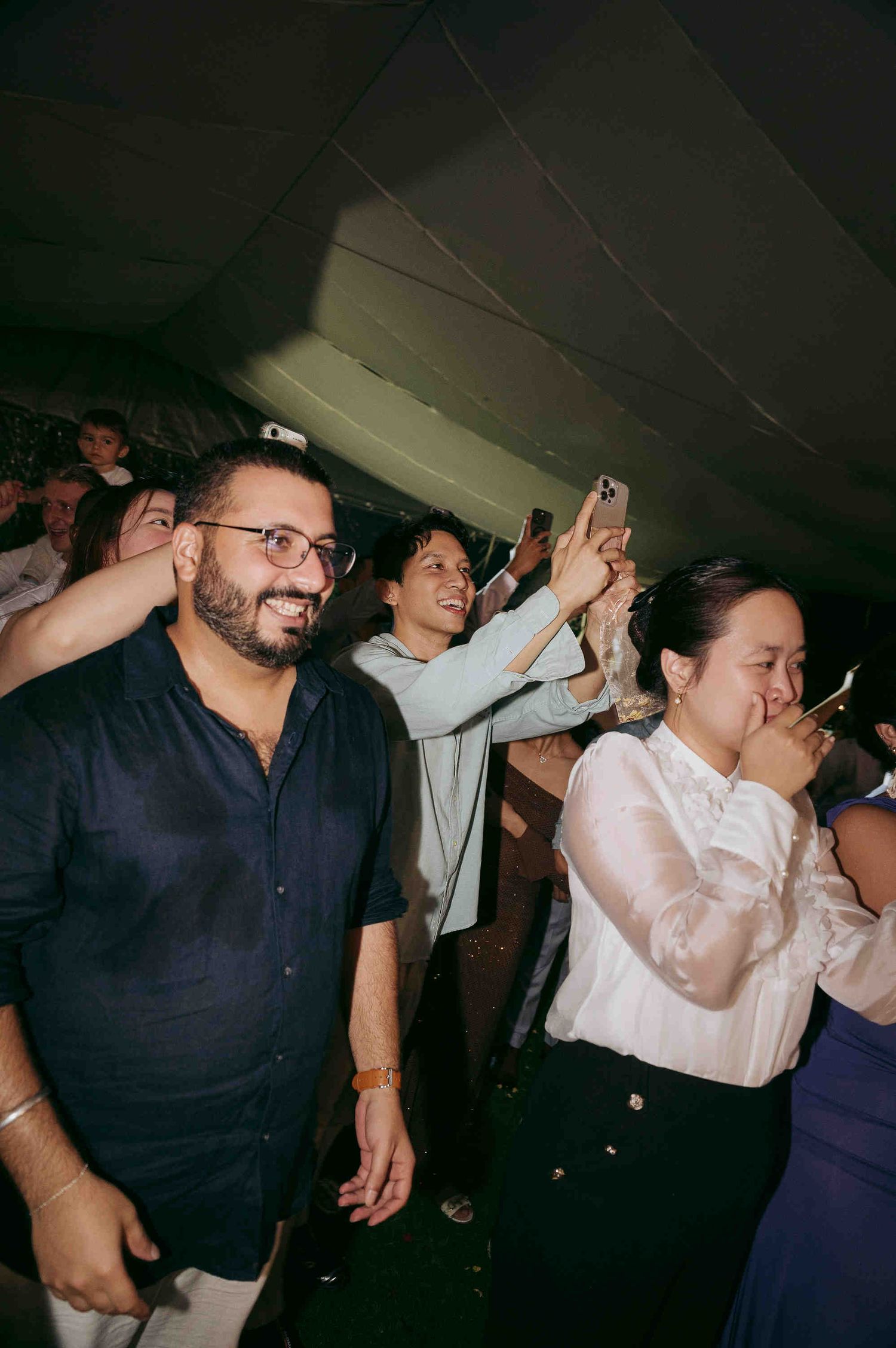 Guests laughing and celebrating at an indoor party event, man in navy shirt smiling, woman in white blouse emotional.