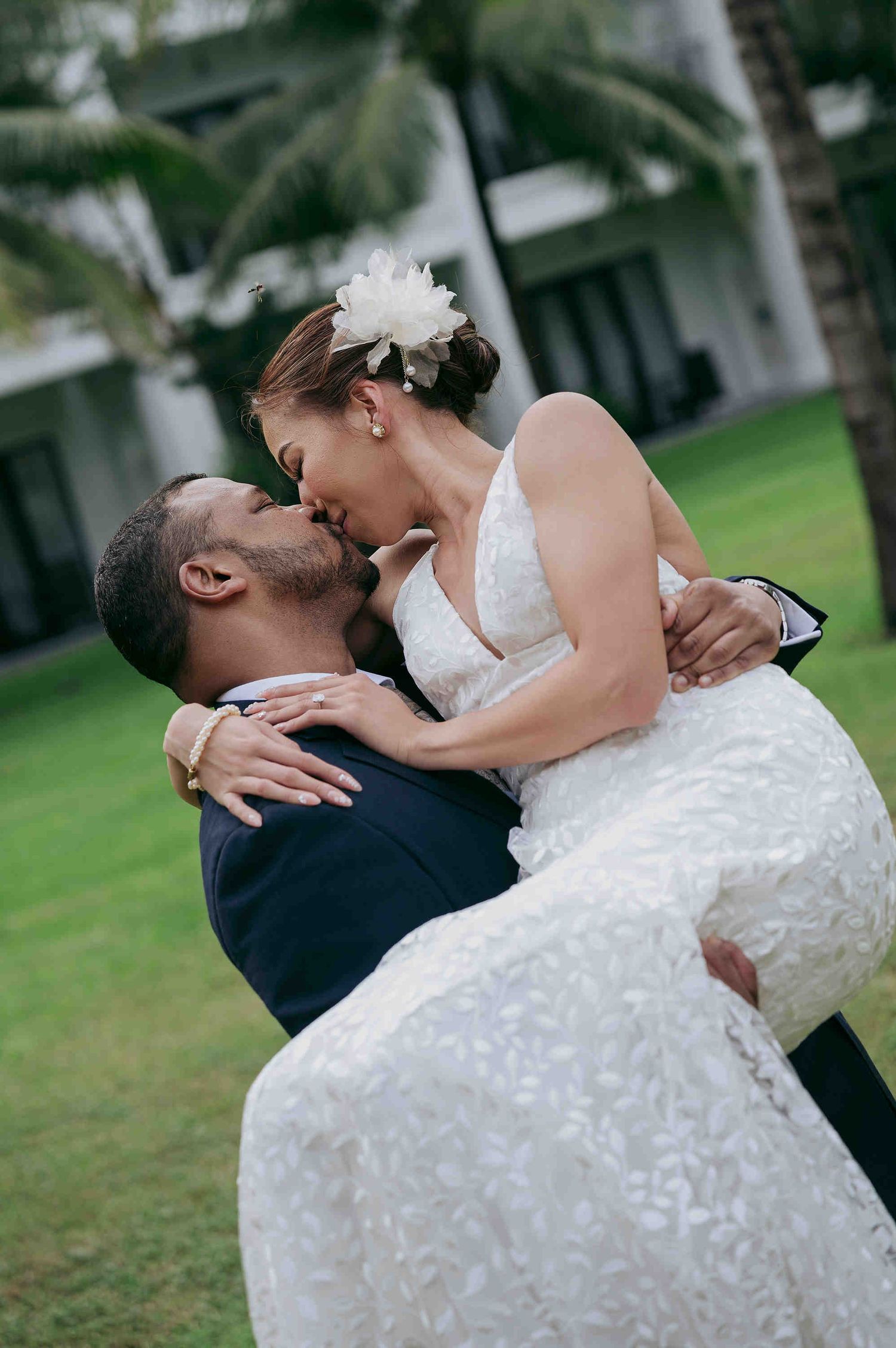 Groom lifts bride in white lace gown for a romantic kiss outdoors with palm trees and green lawn.